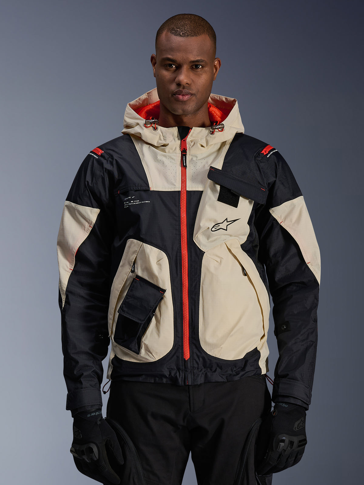 Alpinestars veste Mogress Air, veste de moto urbaine pour homme, amande et noire avec détails orange vif, textile ripstop avec capuche et poches utilitaires, look streetwear porté par un mannequin.