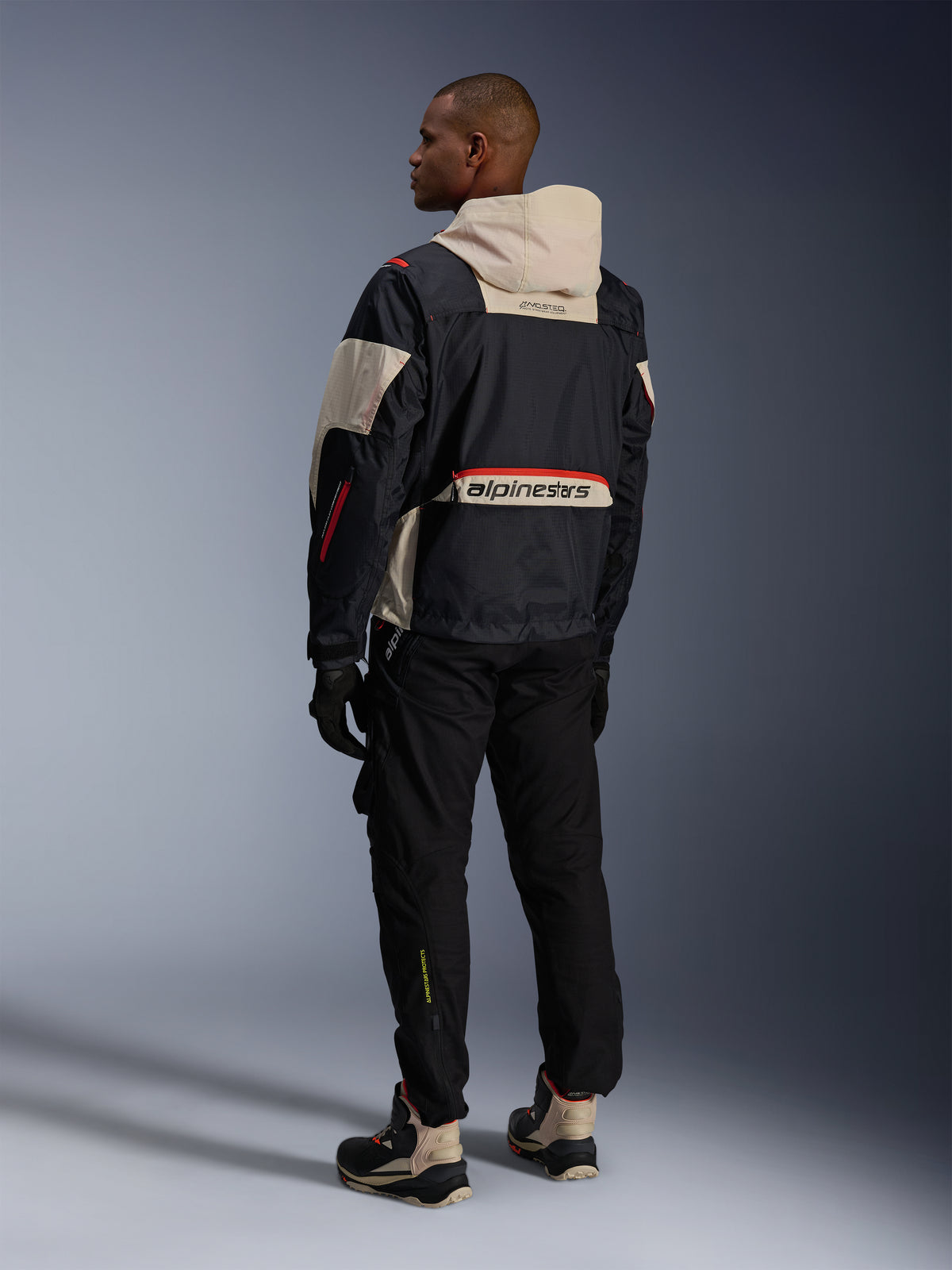 Rear view of a man wearing Alpinestars Acteon Pants in Black, paired with a Mogress Air Jacket in black and cream, black gloves, and riding boots, contemporary urban motorcycle outfit