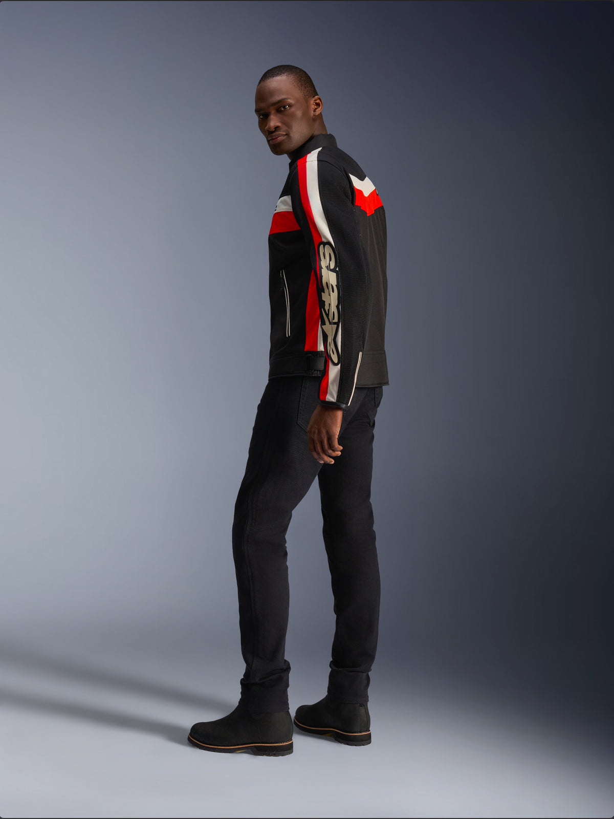 Un homme portant la veste Alpinestars T-Dyno Air en noir, rouge vif et écru, veste de moto en textile d'inspiration vintage avec des panneaux en maille, logo imposant sur la manche, associée à un pantalon et des bottes noirs pour un look motard moderne