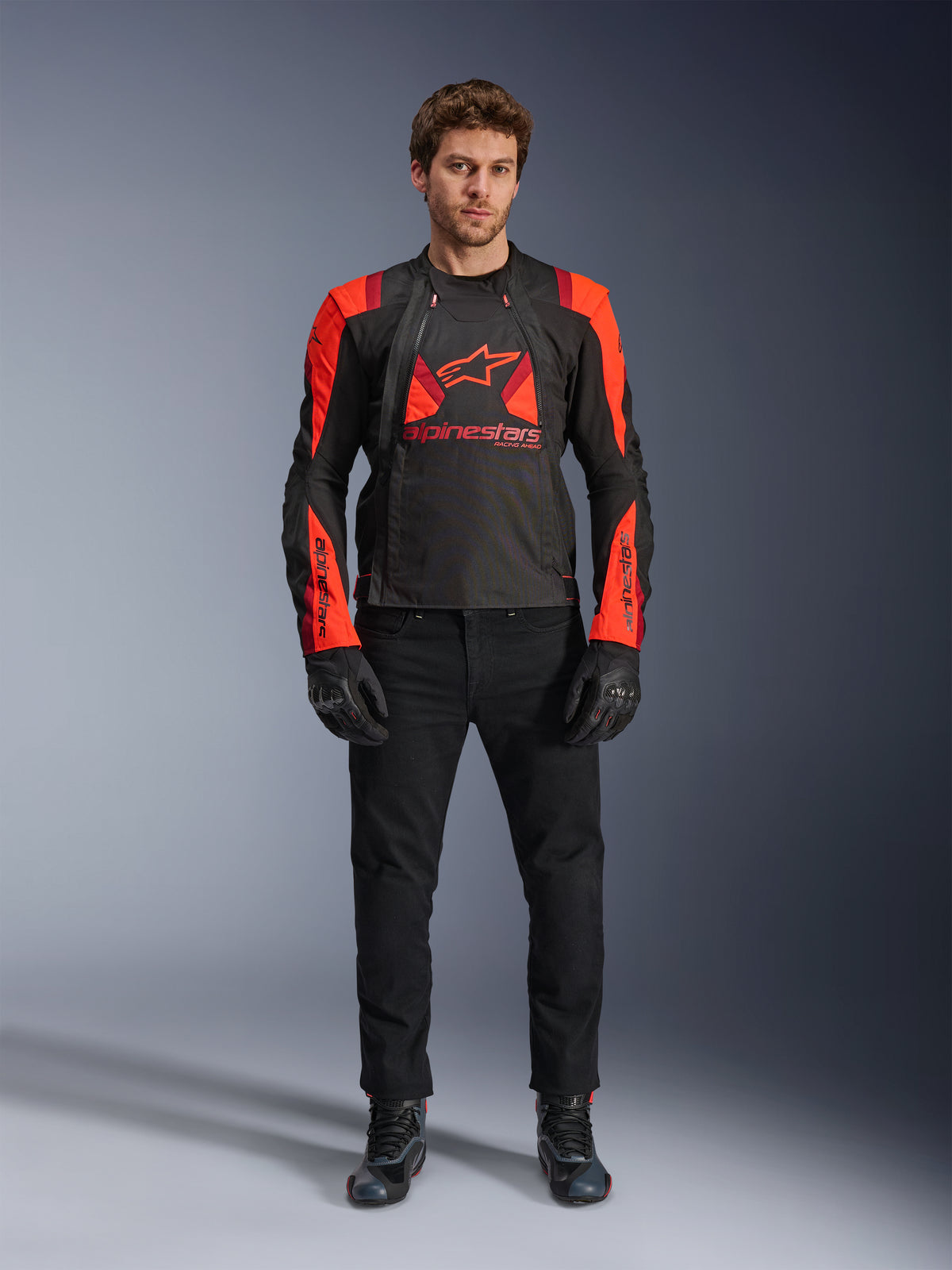 Veste Alpinestars T-Stunt Air, noir, rouge fluo et rouge foncé, portée par un mannequin avec un jean de moto noir et des gants, dotée d'une conception modulaire 2-en-1 avec des panneaux en mesh pour la circulation de l'air, équipement de moto à coupe sportive pour la route