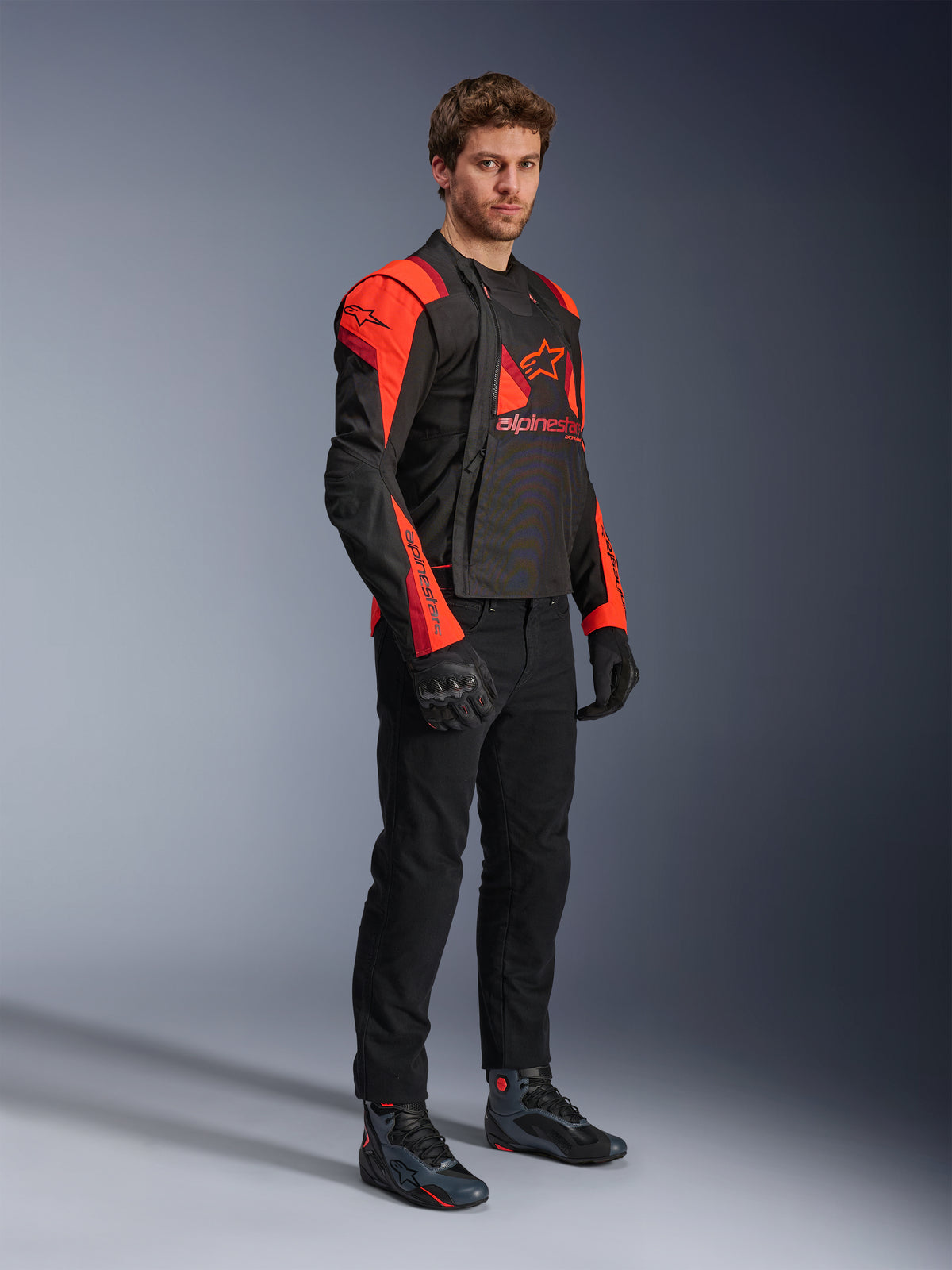 Veste Alpinestars T-Stunt Air, noir, rouge fluo et rouge foncé, veste de moto modulaire portée par un mannequin avec les panneaux avant ouverts pour révéler l'intérieur en maille, accompagnée d'un pantalon, de gants et de chaussures de moto noirs.