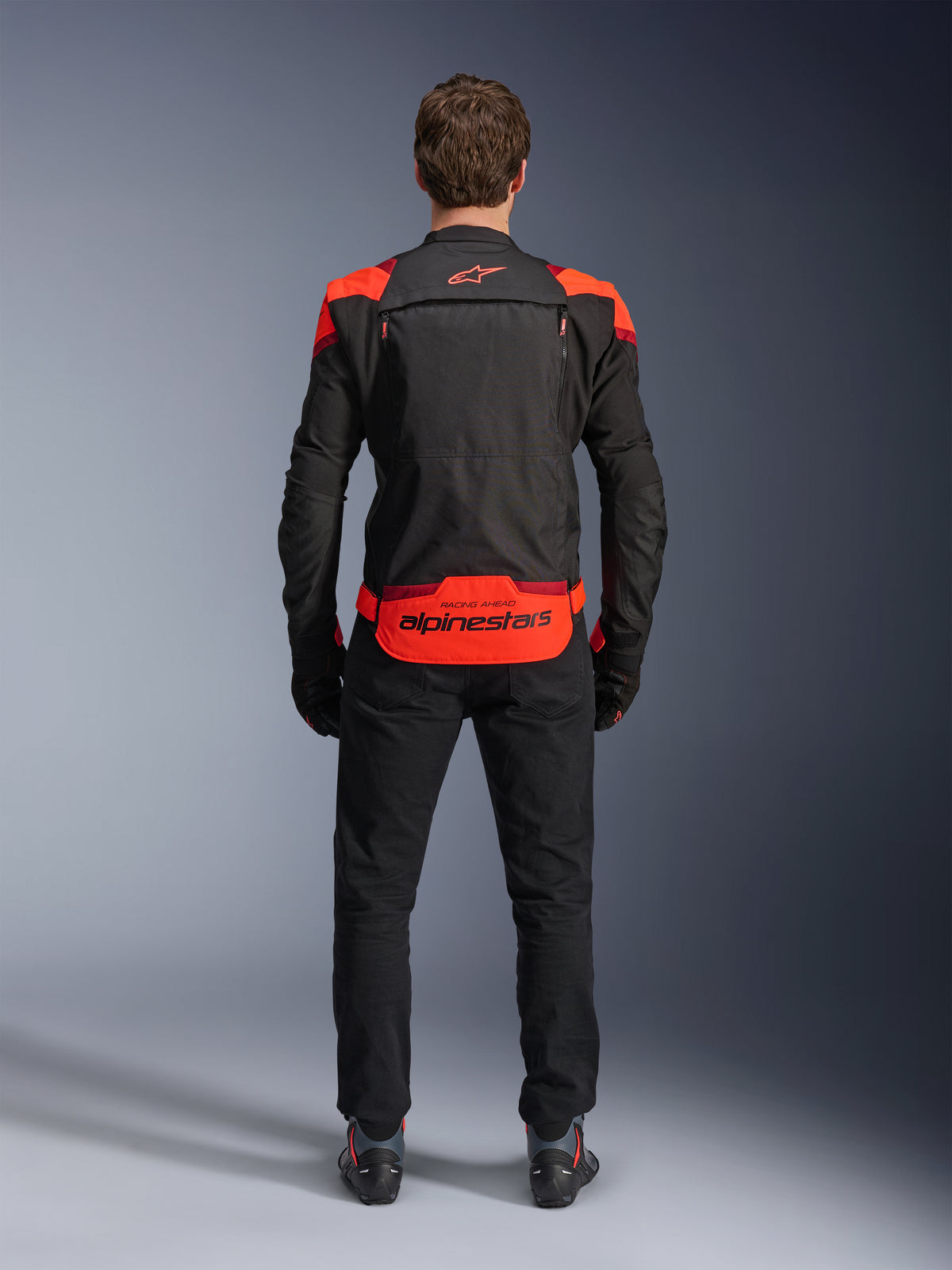 Veste Alpinestars T-Stunt Air, noire avec accents Rouge Fluo et Rouge Foncé, vue de dos sur modèle, accompagnée d'un pantalon de moto et de bottes noirs, arborant un marquage Alpinestars imposant sur le bas du dos, conception modulaire avec mesh pour la ventilation.