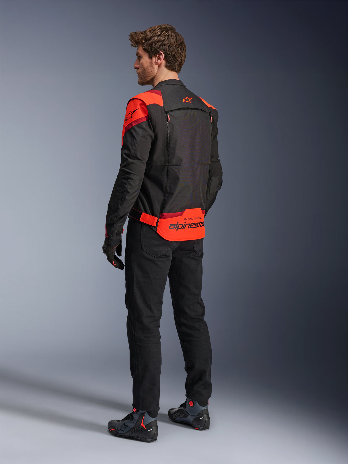 Homme portant la veste Alpinestars T-Stunt Air vue de dos, Noir Rouge Fluo et Rouge Foncé, veste de moto en textile avec panneaux en mesh et coupe sportive, avec le logo Alpinestars imposant dans le bas du dos, associée à un pantalon noir et des bottes de moto