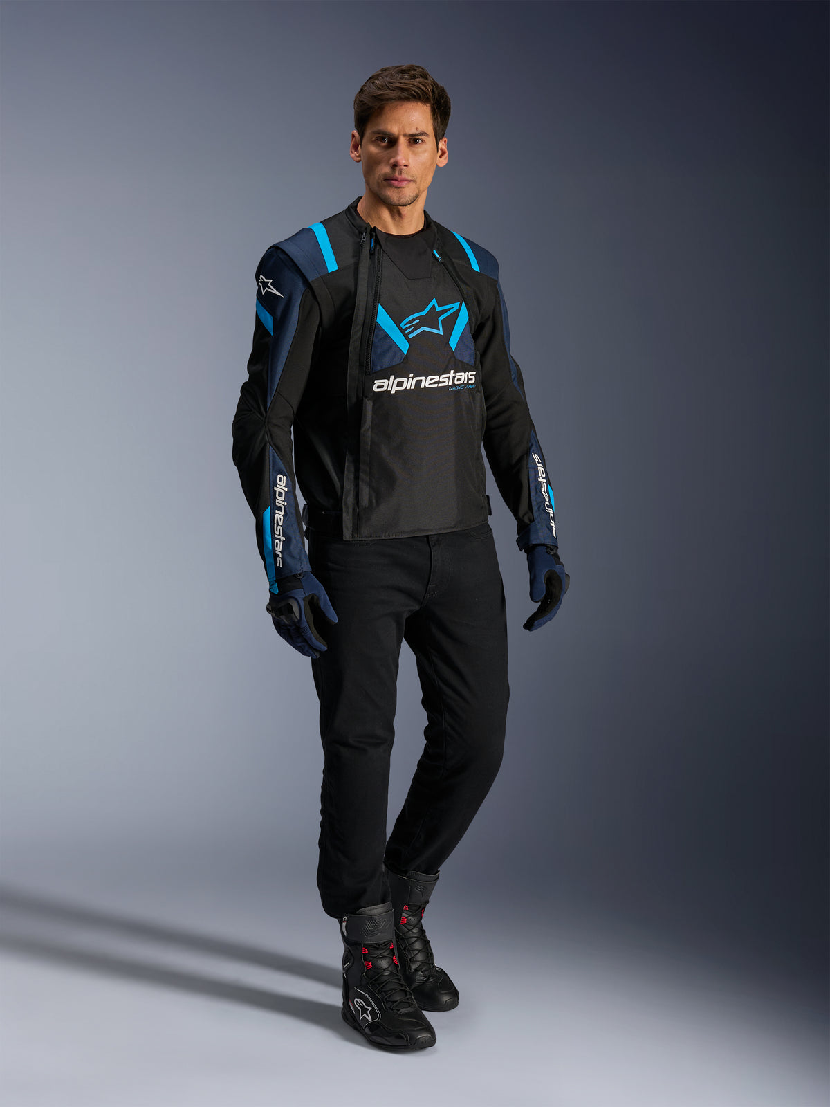 Modèle masculin portant la veste Alpinestars T-Stunt Air, noir, bleu foncé et bleu vif, dotée de larges panneaux en mesh pour la circulation de l'air, associée à un pantalon noir et des gants de moto, veste de moto à coupe sportive pour la route en été.