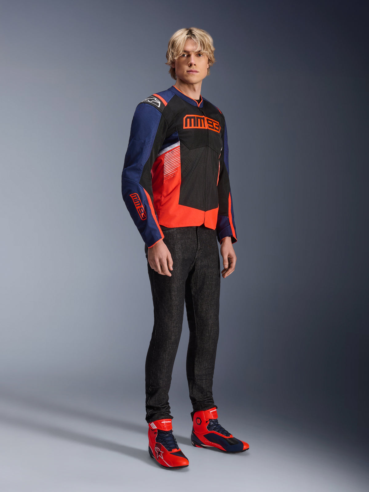 Male model wearing the Alpinestars MM93 T-SPS Air V2 Jacket in Blue Navy, Black, and Red Fluo, featuring bold MM93 branding on the chest and sleeves, paired with dark jeans and matching red and blue motorcycle boots, sporty urban riding gear