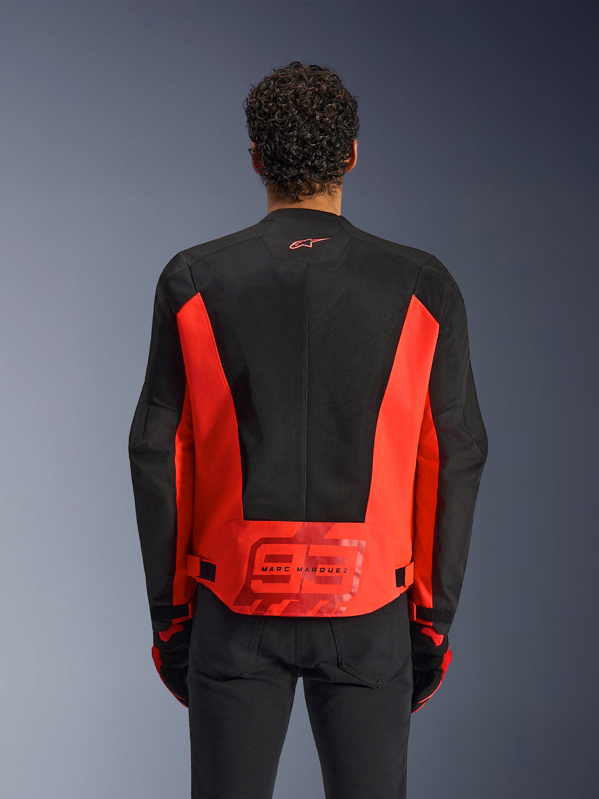 Vue arrière d'un homme portant la veste Alpinestars MM93 C-1 Air en rouge fluo et noir, arborant un grand logo « 93 » Marc Marquez au bas du dos, des panneaux en maille légère pour la ventilation, associée à un pantalon et des gants noirs.