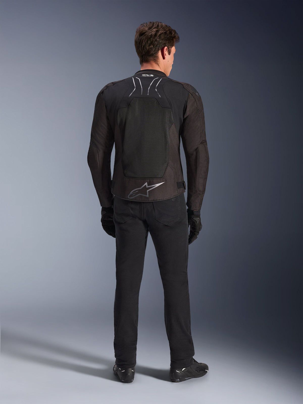 Veste Alpinestars SMX Air, Noir Anthracite, vue de dos sur modèle masculin, avec un grand logo Alpinestars argenté dans le bas du dos et des empiècements en maille respirante, portée avec un pantalon de moto, des gants et des bottes noirs, équipement moto sportif