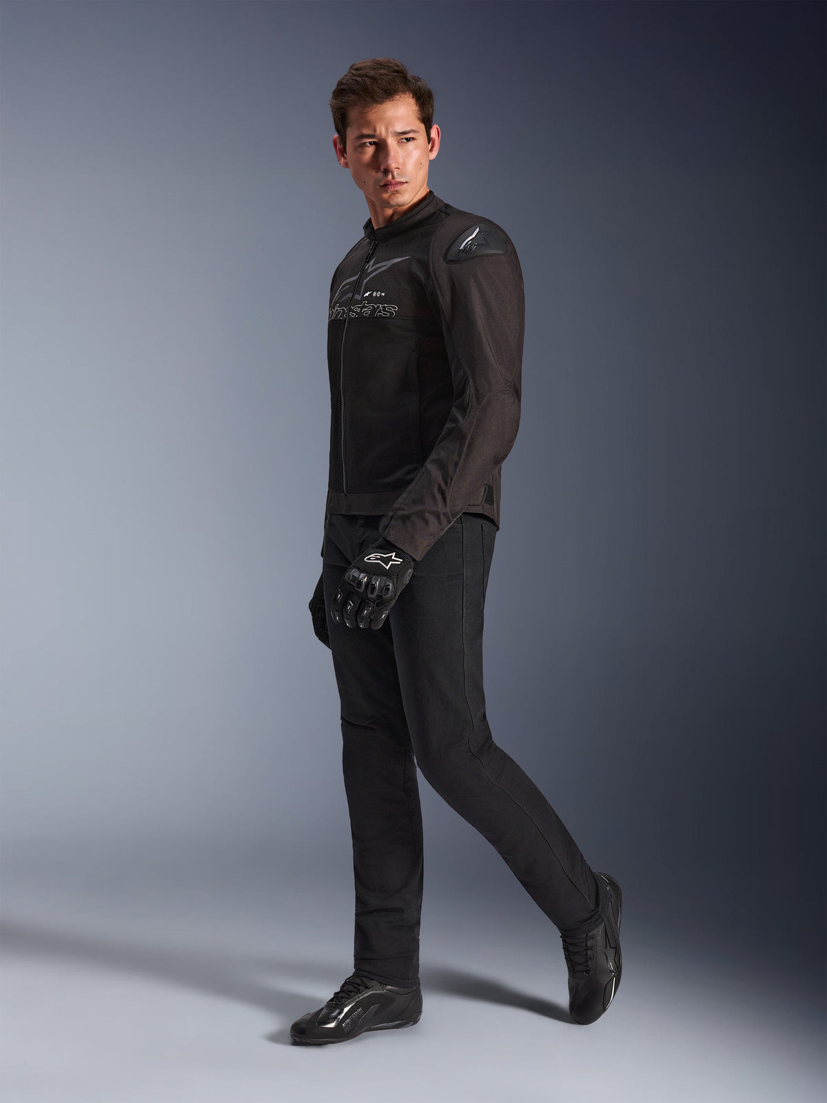Veste Alpinestars SMX Air en Noir Anthracite, portée par un mannequin avec pantalon de moto, gants et chaussures noirs assortis, cliché studio en pied, équipement moto sport de performance avec maille technique et sliders d'épaule