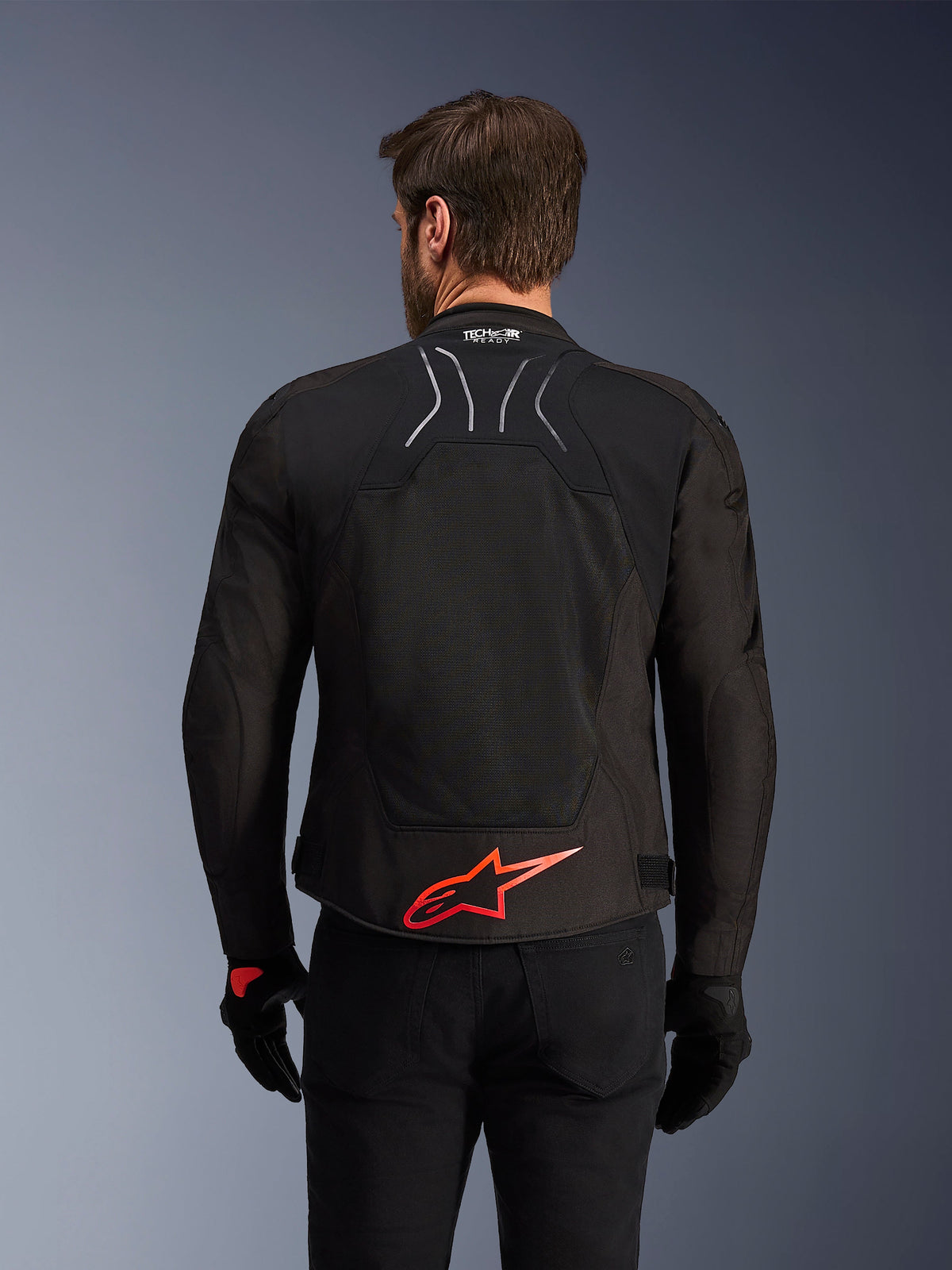 Veste Alpinestars SMX Air, vue arrière, noire avec logo Alpinestars rouge vif dans le bas du dos, dotée d'un panneau en maille technique à haute circulation d'air et du marquage Tech-Air Ready, portée par un pilote avec pantalon et gants noirs