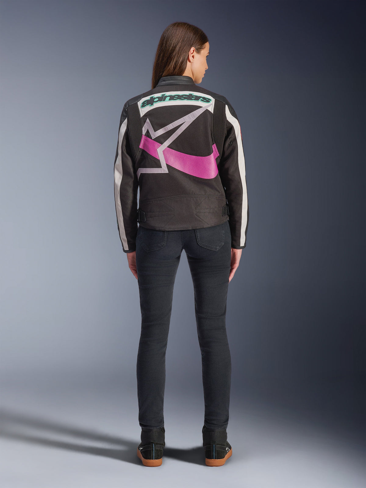 Vue arrière d'une femme portant la veste Idles d'Alpinestars, noire avec des rayures blanc cassé, ornée d'un grand logo étoile Alpinestars violet et blanc et d'un lettrage vert au dos, associée à un jean slim foncé, vêtement de moto urbain d'inspiration vintage
