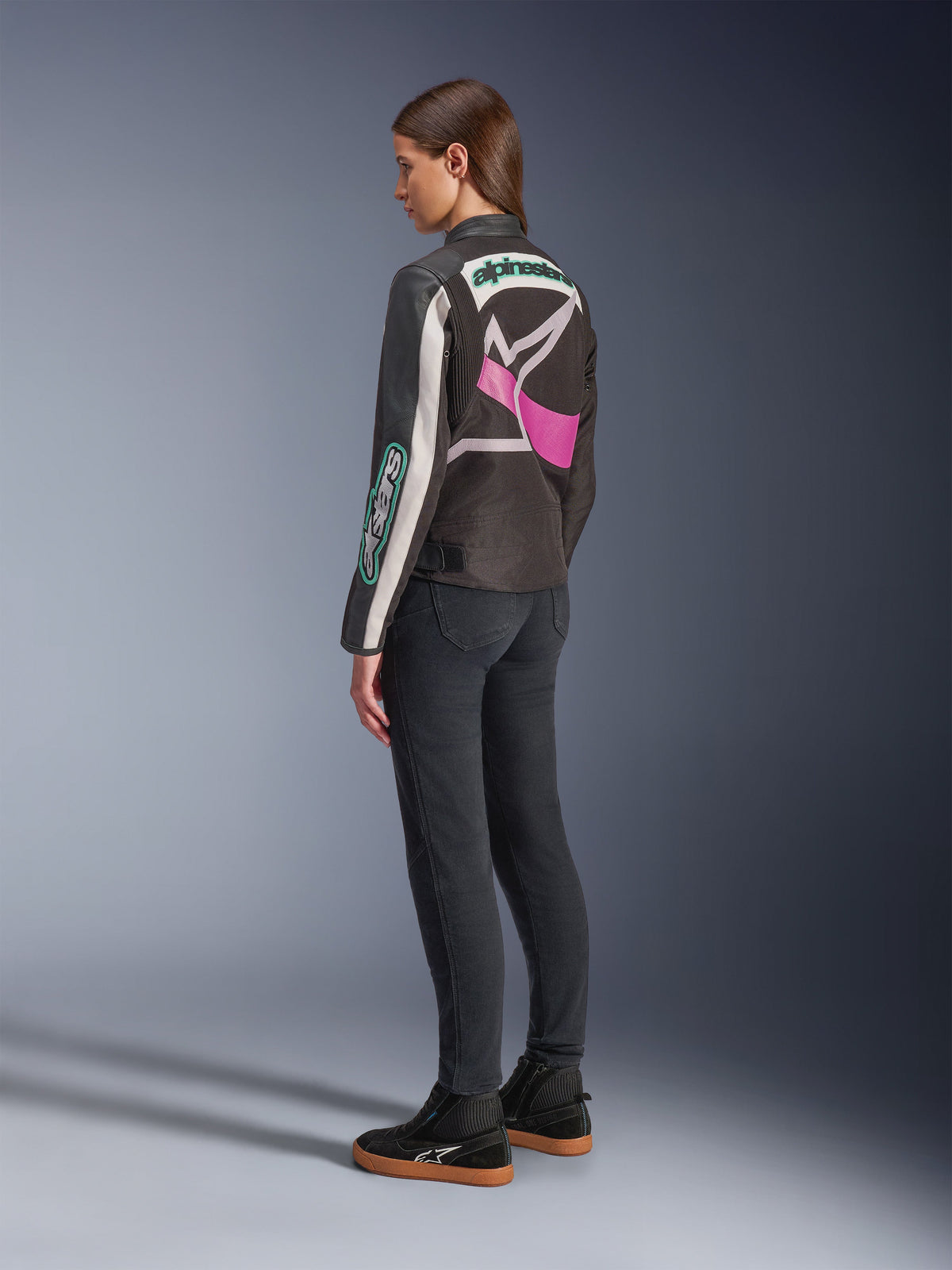 Veste femme Idles Alpinestars, veste de moto, noir, blanc cassé et violet, vue de dos sur mannequin, présentant un grand logo Alpinestars enveloppant et un design inspiré de la course vintage, portée avec un jean foncé et des chaussures noires.