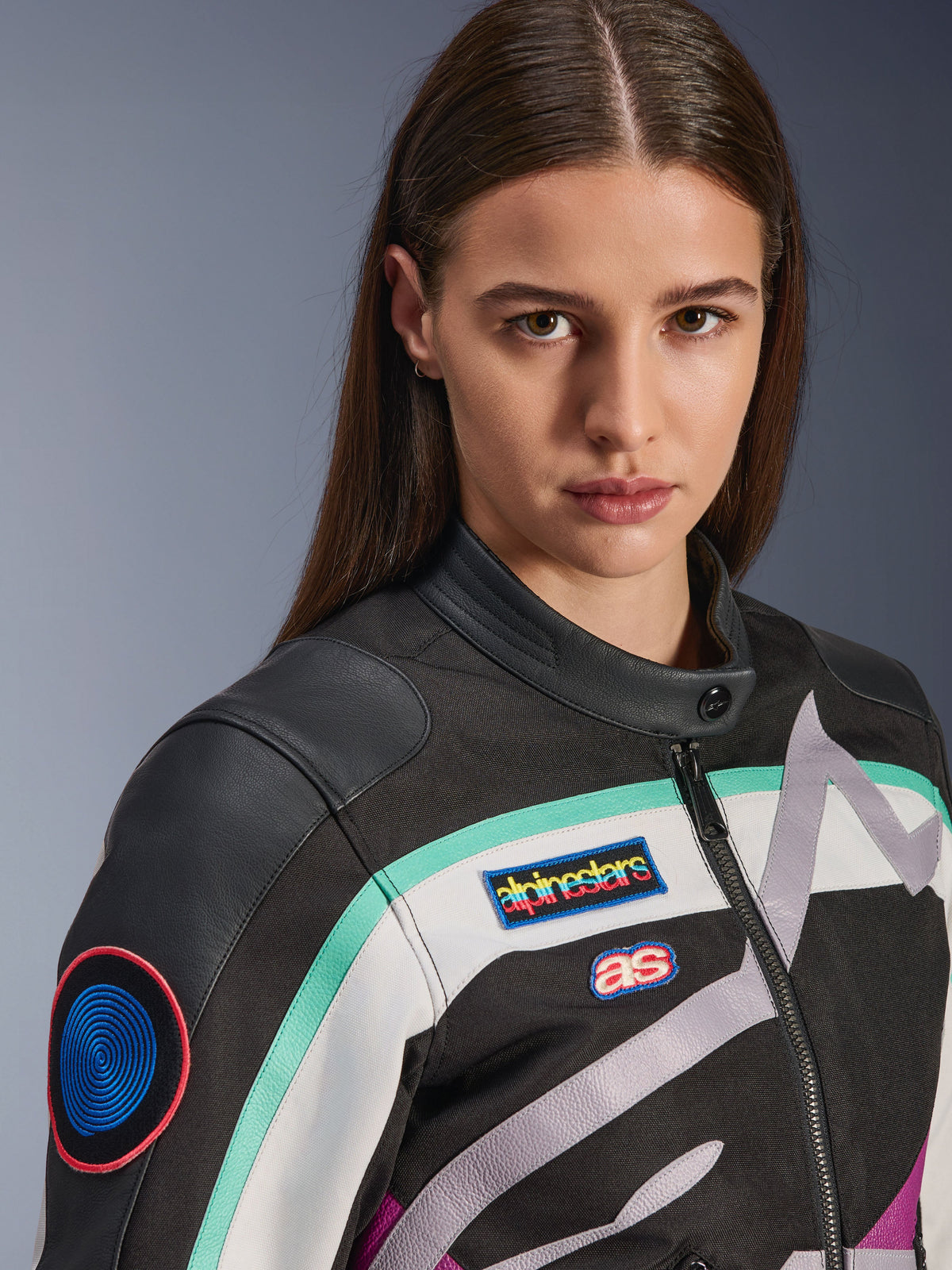 Veste Alpinestars Idles pour femme, veste de moto, noir, blanc cassé et violet avec des touches de bleu sarcelle, gros plan mettant en valeur les écussons héritage Alpinestars colorés et les détails d'épaule en cuir de chèvre, design d'inspiration vintage pour la conduite urbaine.