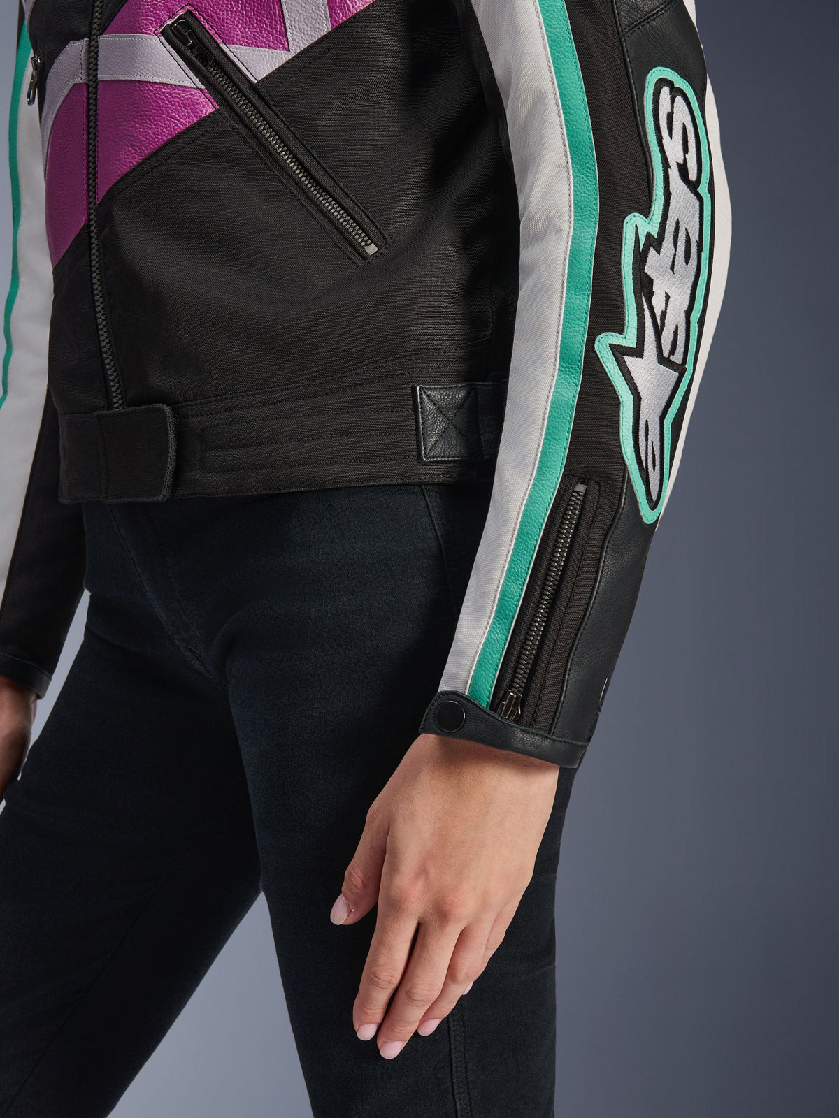 Veste femme Alpinestars Idles, vue rapprochée de la manche et du côté du torse, noire avec accents violets, blancs et bleu sarcelle, logo Alpinestars imposant sur le bras, poignet zippé avec bouton-pression et sangle de réglage à la taille, équipement de moto d'inspiration vintage
