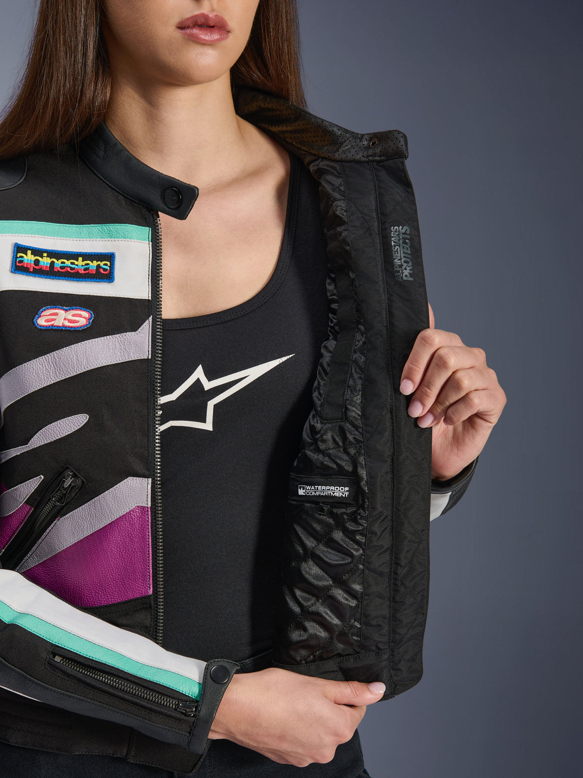 Veste Idles Femme Alpinestars, Noir Blanc Cassé Violet, vue rapprochée montrant la doublure matelassée interne avec un compartiment imperméable, ornée d'écussons de course d'inspiration vintage et d'accents colorés bleu sarcelle et violet sur l'extérieur en textile.