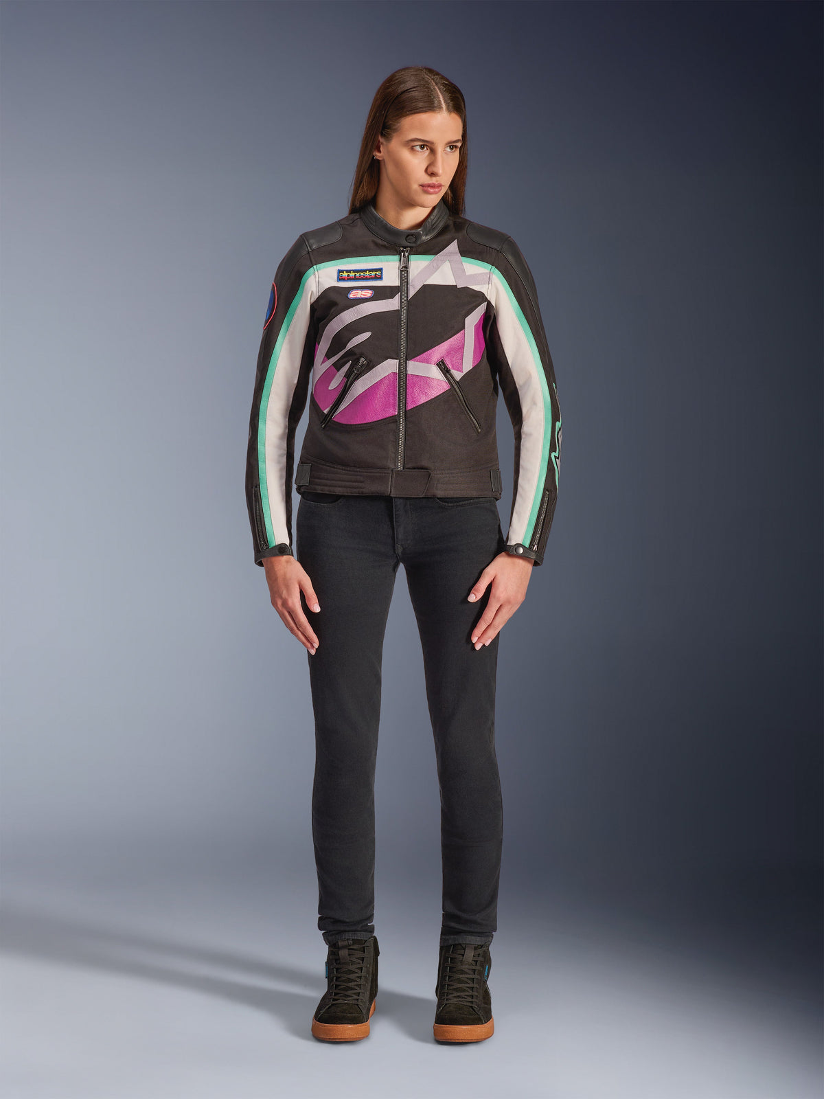 Femme portant la veste Alpinestars Idles pour femme, noir, blanc cassé et violet, design d'inspiration racing vintage avec un grand logo enveloppant et des accents bleu sarcelle, assortie d'un jean noir et de baskets de moto, vêtement de moto urbain