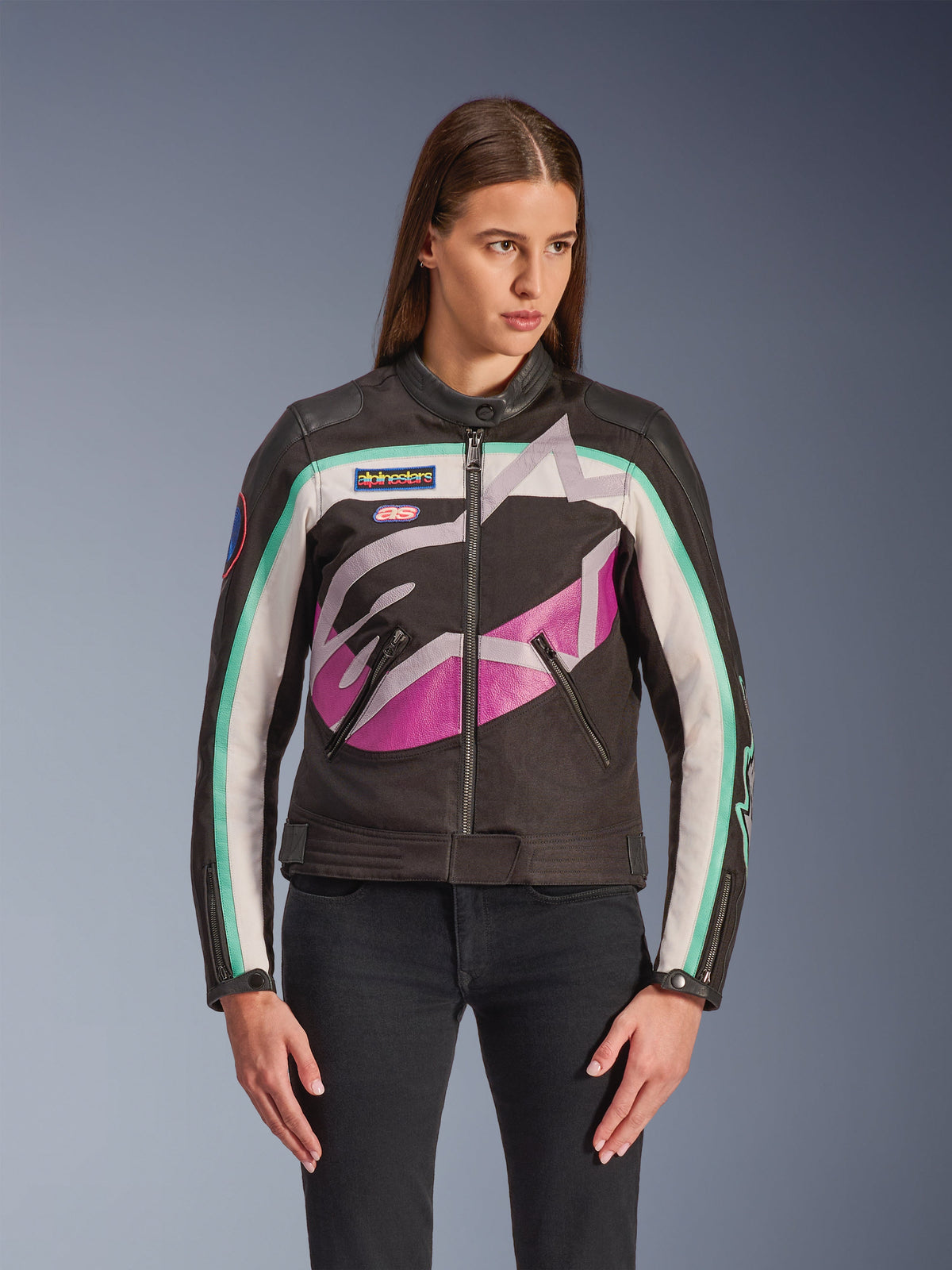 Veste pour femme Alpinestars Idles, veste de moto, noire avec design color-block blanc cassé, violet et turquoise, style vintage inspiré de la course avec grand logo abstrait sur la poitrine, écussons de la marque et fermeture éclair frontale, portée par un mannequin avec un jean noir
