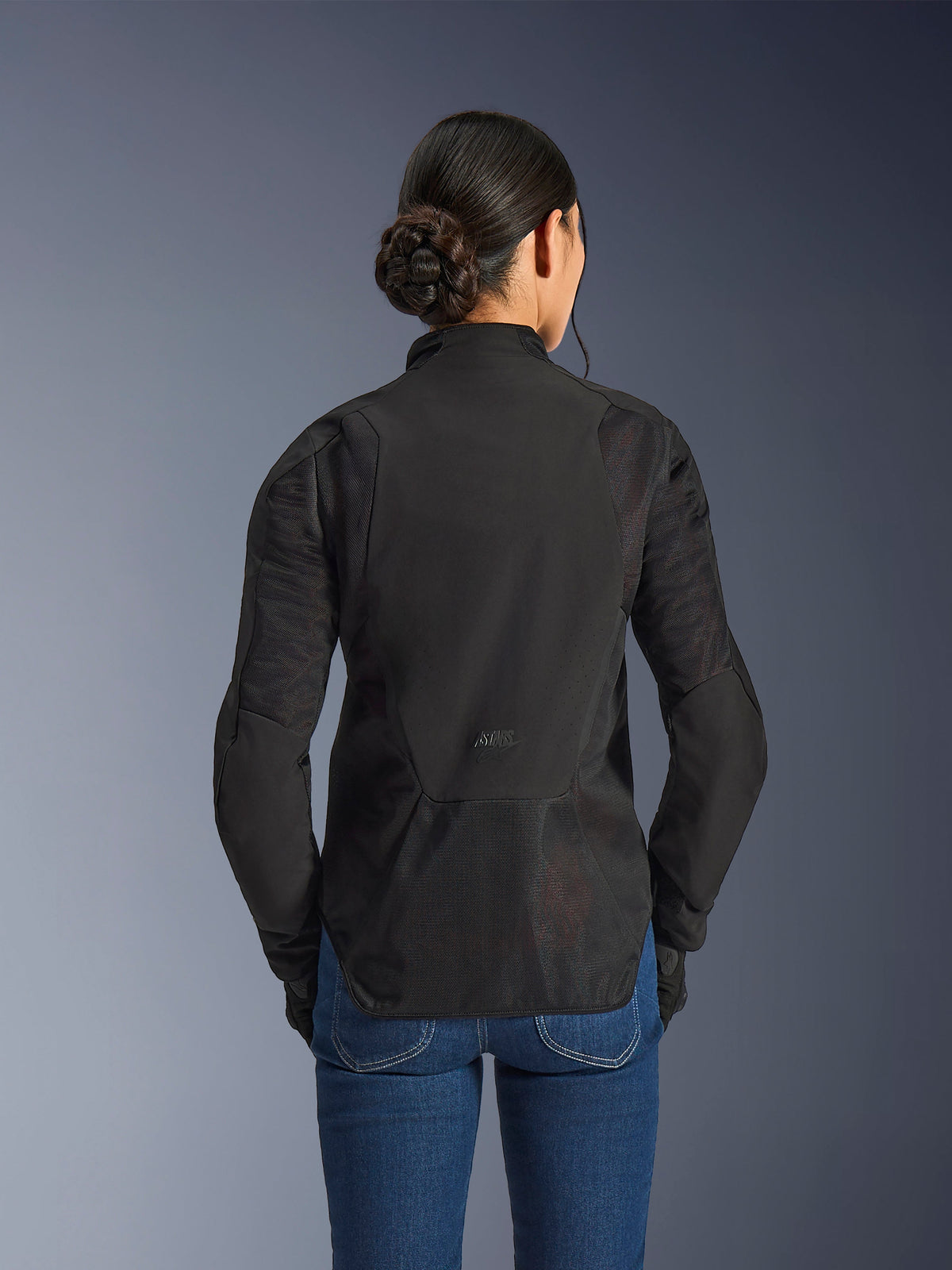 Veste Alpinestars Stella Aeroshell Airflow pour femme, Noir Anthracite, vue arrière montrant les panneaux de ventilation en maille intégrés sur le dos et les manches, châssis en softshell perforé au laser avec un profil arrière allongé, portée avec un jean en denim bleu.