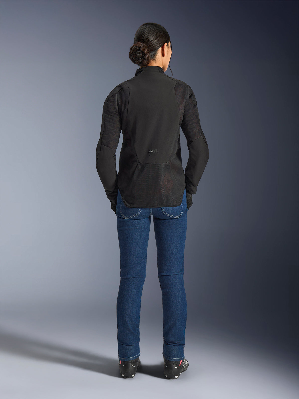 Veste Stella Aeroshell Airflow Alpinestars, Noir Anthracite, vue de dos sur un mannequin, dotée d'un châssis softshell léger avec panneaux en maille perforés au laser, profil arrière allongé pour une meilleure couverture, coupe sport, portée avec un jean bleu