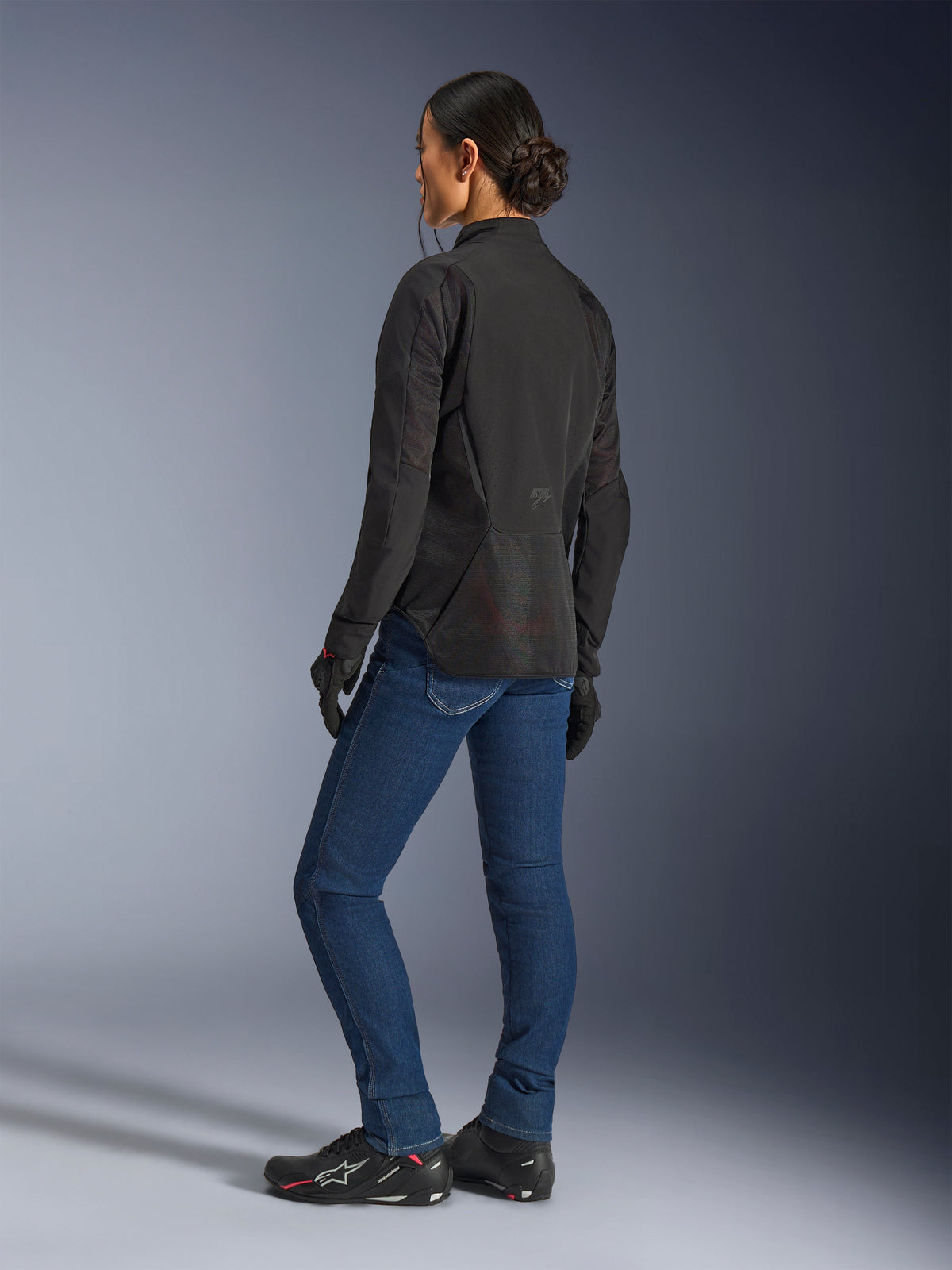 Femme portant la veste Alpinestars Stella Aeroshell Airflow, Noir Anthracite, vue arrière de trois-quarts montrant les panneaux en maille respirante et le profil arrière allongé, associée à un jean de moto en denim bleu et des chaussures Alpinestars noires