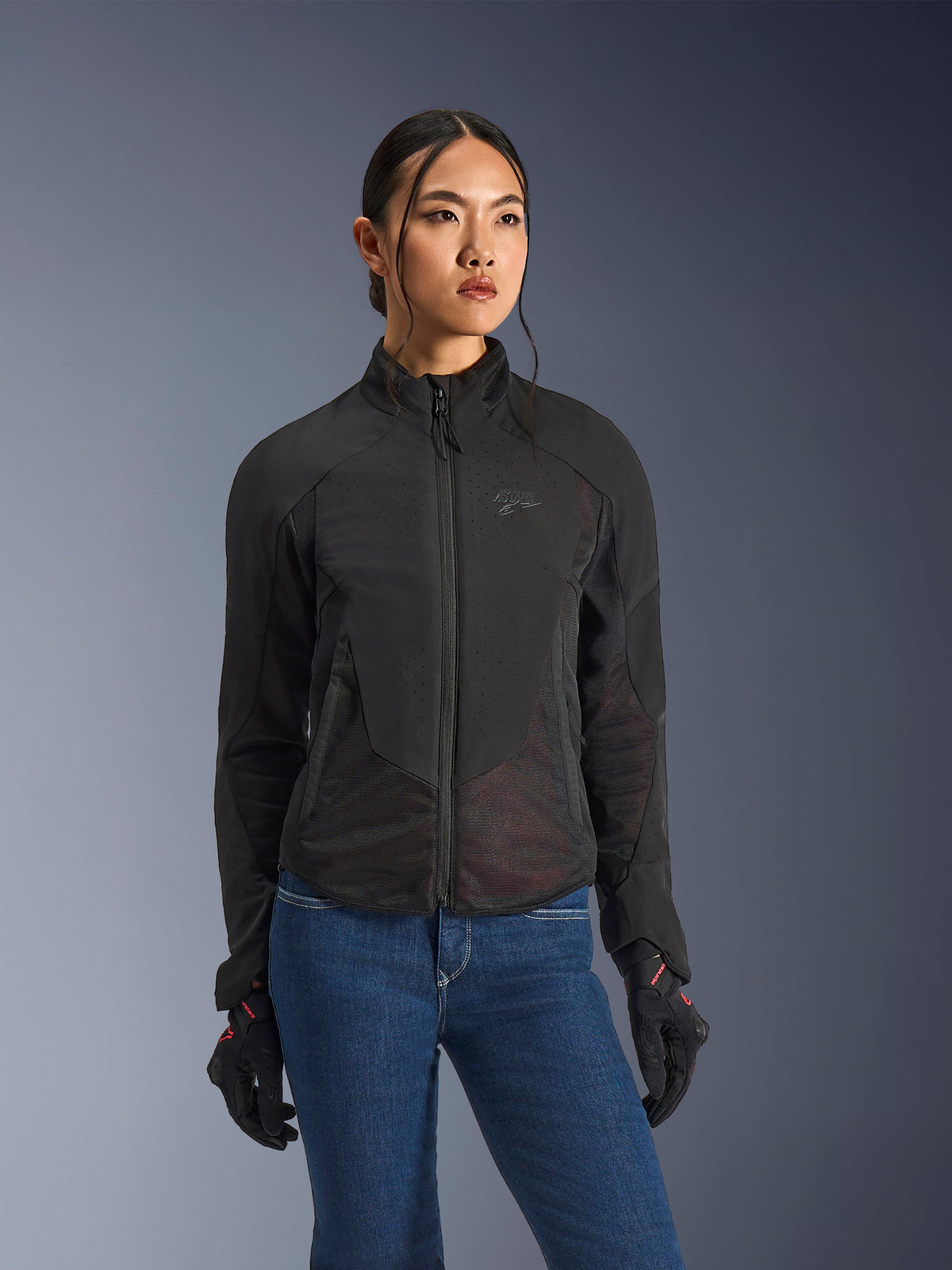 Veste Alpinestars Stella Aeroshell Airflow, veste de moto pour femme, noir anthracite, portée par un mannequin avec un jean bleu et des gants noirs, dotée d'un softshell perforé au laser et de panneaux en maille pour une ventilation optimale, design ergonomique pour la conduite sportive.