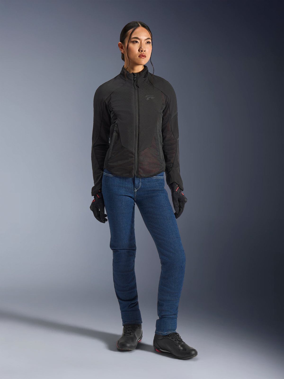 Motarde portant la veste Alpinestars Stella Aeroshell Airflow en noir, avec ventilation perforée au laser et panneaux en maille, associée à un jean de moto bleu et un équipement de moto noir, esthétique urbaine et sportive moderne