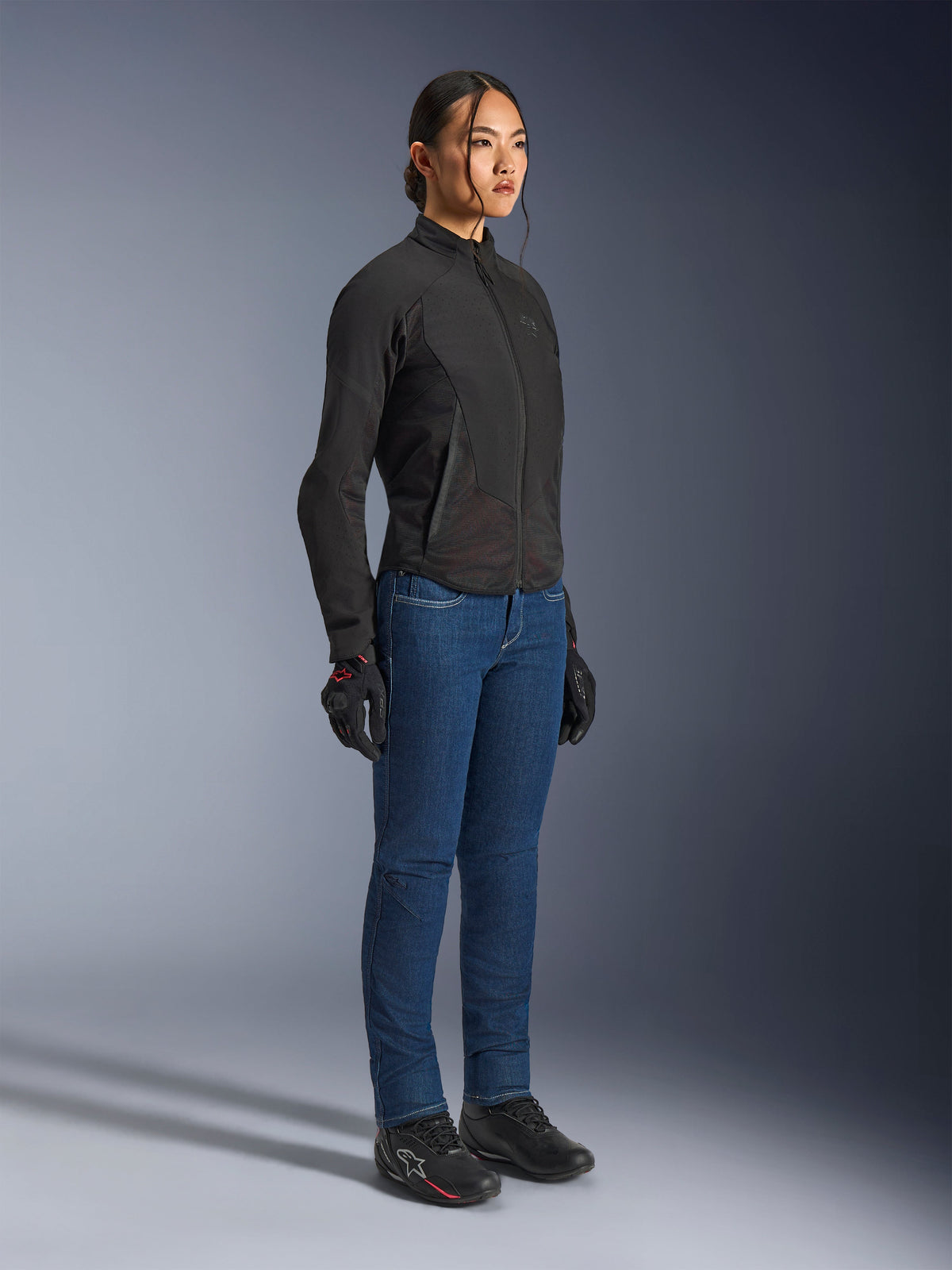 Motarde portant la veste Alpinestars Stella Aeroshell Airflow en Black Anthracite, veste de moto softshell à coupe ajustée, associée à un jean de moto bleu et des gants noirs, vue studio en pied