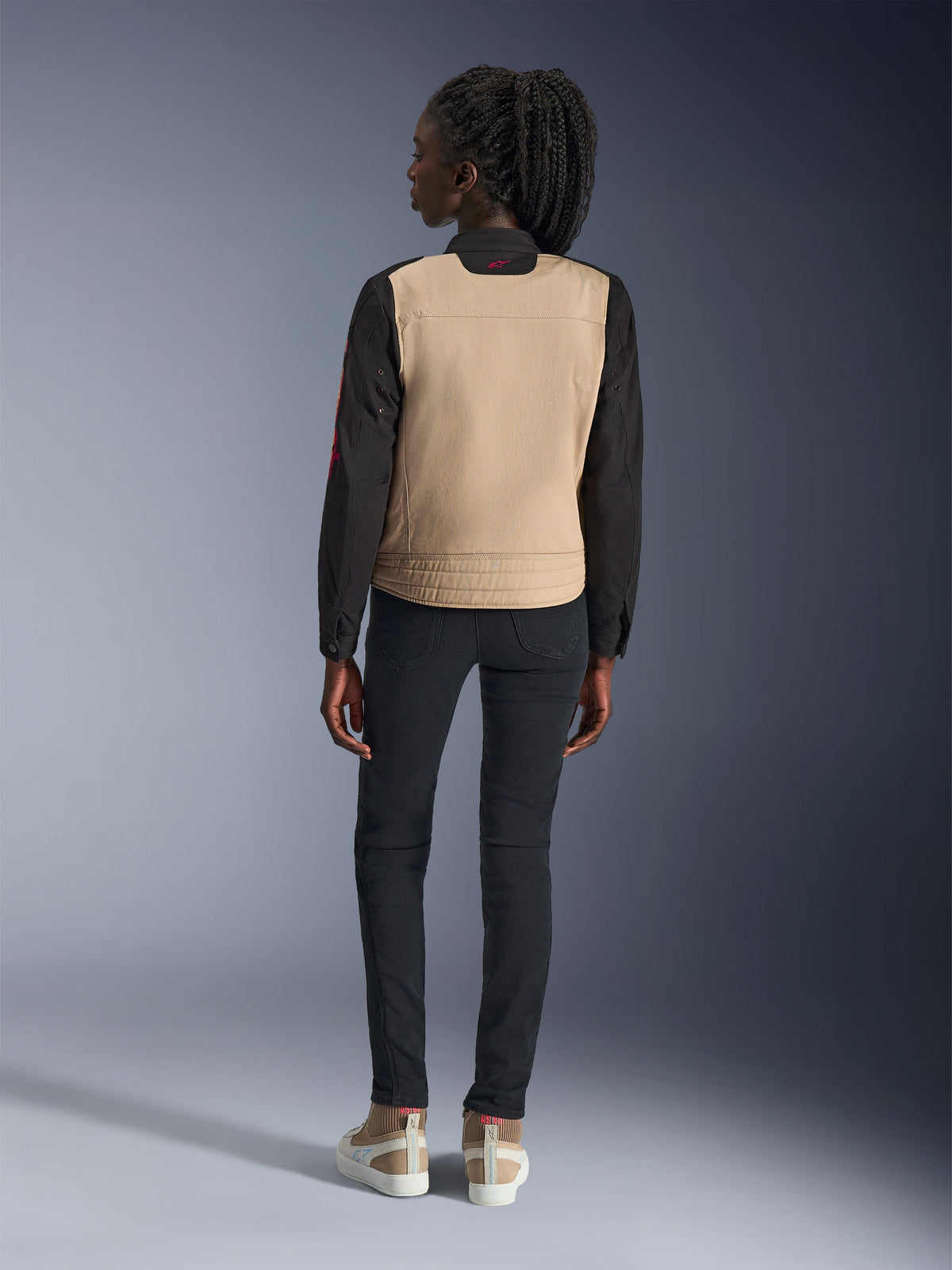 Femme portant la veste Alpinestars Stella Grace, Aluminum Black avec torse beige et manches noires, vue de dos, associée à un pantalon slim noir et des baskets décontractées, équipement de moto urbain pour femme