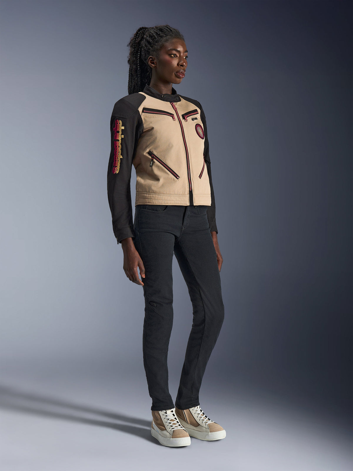Modèle portant la veste Alpinestars Stella Grace en Aluminum Black, avec un buste beige et des manches noires aux accents de zips rouges, associée à un pantalon noir ajusté et des baskets crème, équipement de moto urbain contemporain pour femme
