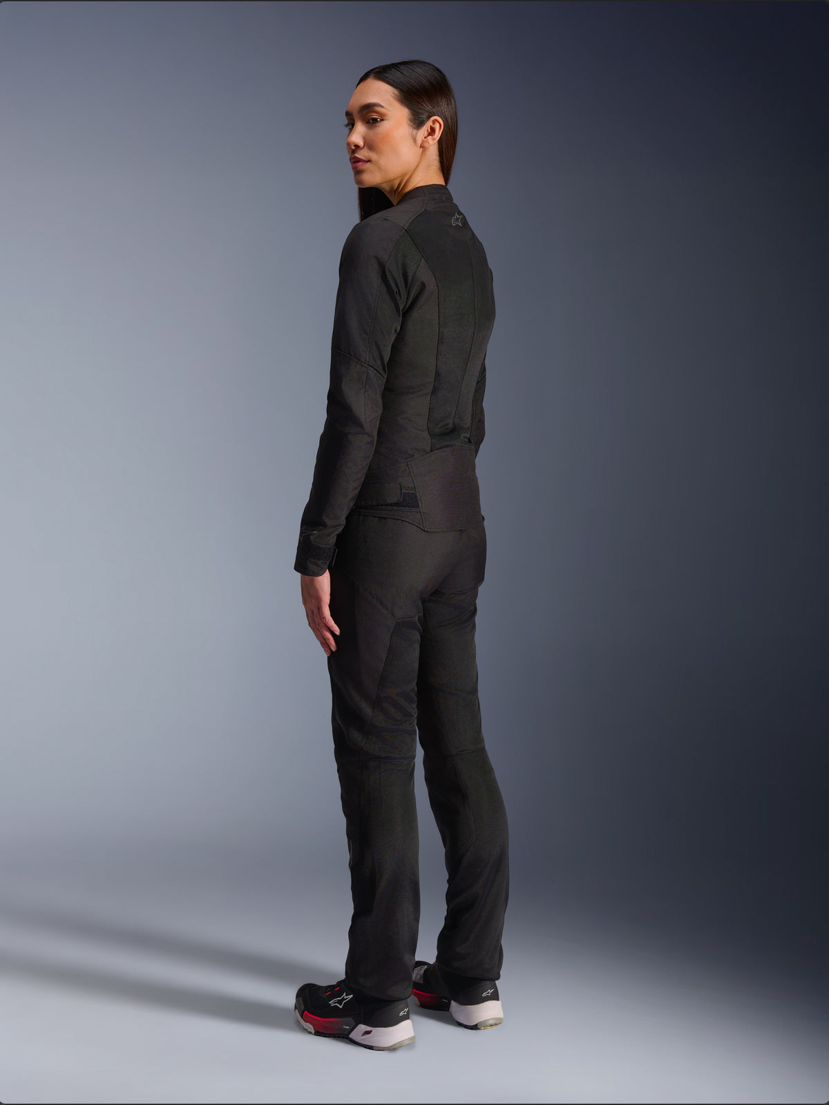 Woman wearing Alpinestars Stella C-1 Air Jacket in Black, urban textile motorcycle jacket with mesh panels, rear-side view showing the sleek fit and upper back logo, worn with matching black pants and athletic shoes, contemporary motorcycle gear.
