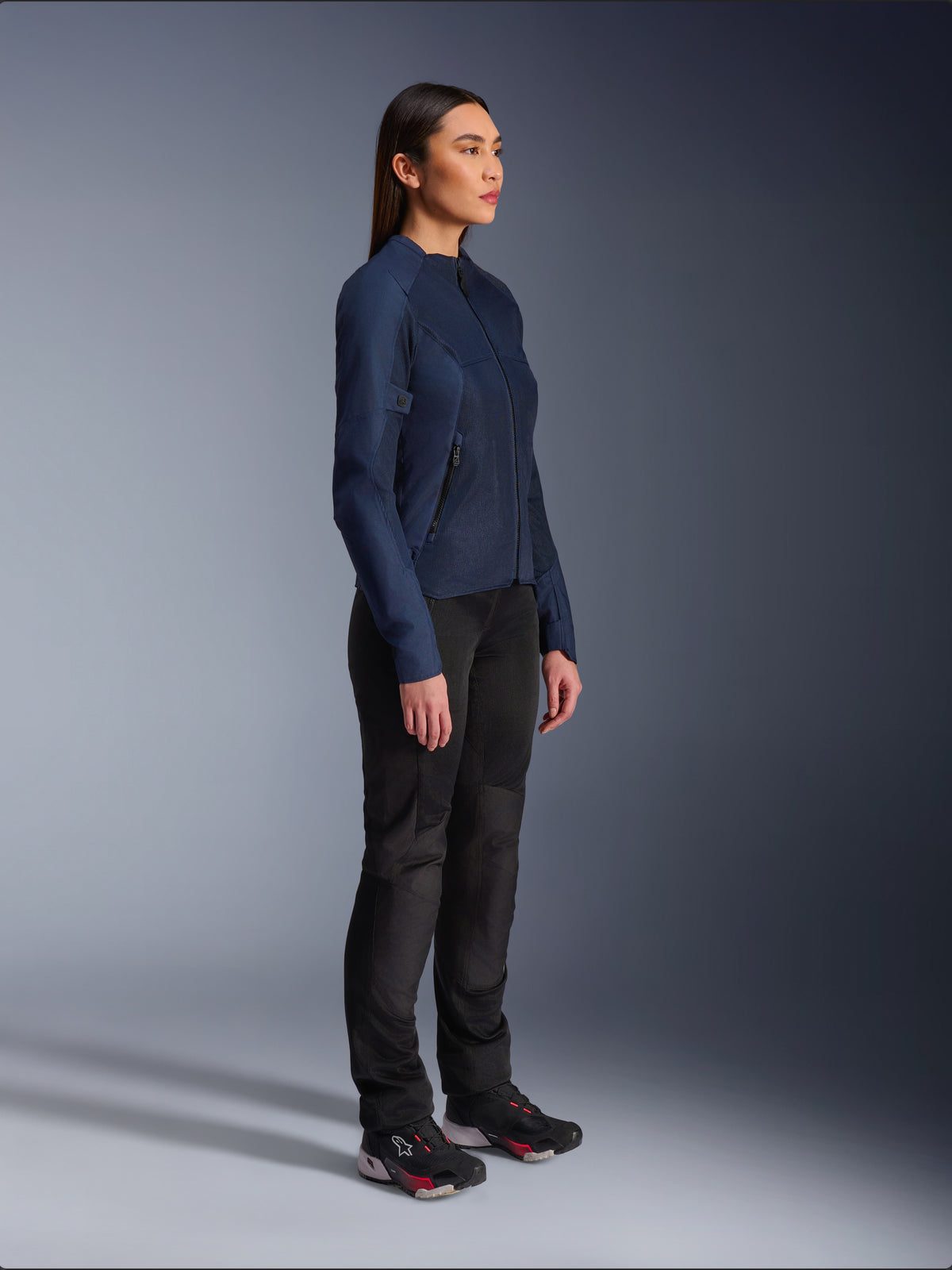 Femme portant la veste Alpinestars Stella C-1 Air en bleu, une veste de moto urbaine avec des panneaux en maille légère pour la ventilation, associée à un pantalon de moto noir et des chaussures techniques, présentant un profil épuré et un marquage raffiné, vue de profil en studio
