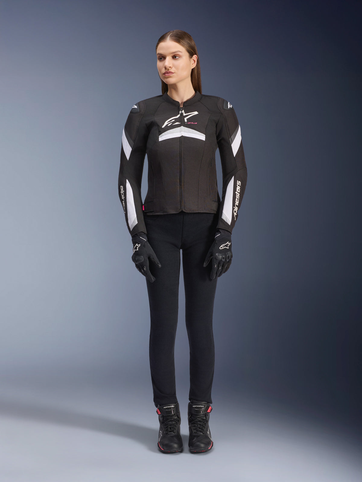 Mannequin femme portant la veste Alpinestars Stella T-GP Plus R V4 en noir et blanc, veste de moto sport en textile, accompagnée d'un pantalon de moto, de gants et de bottes noirs, vue de face montrant le logo Alpinestars blanc et les détails sur les manches.