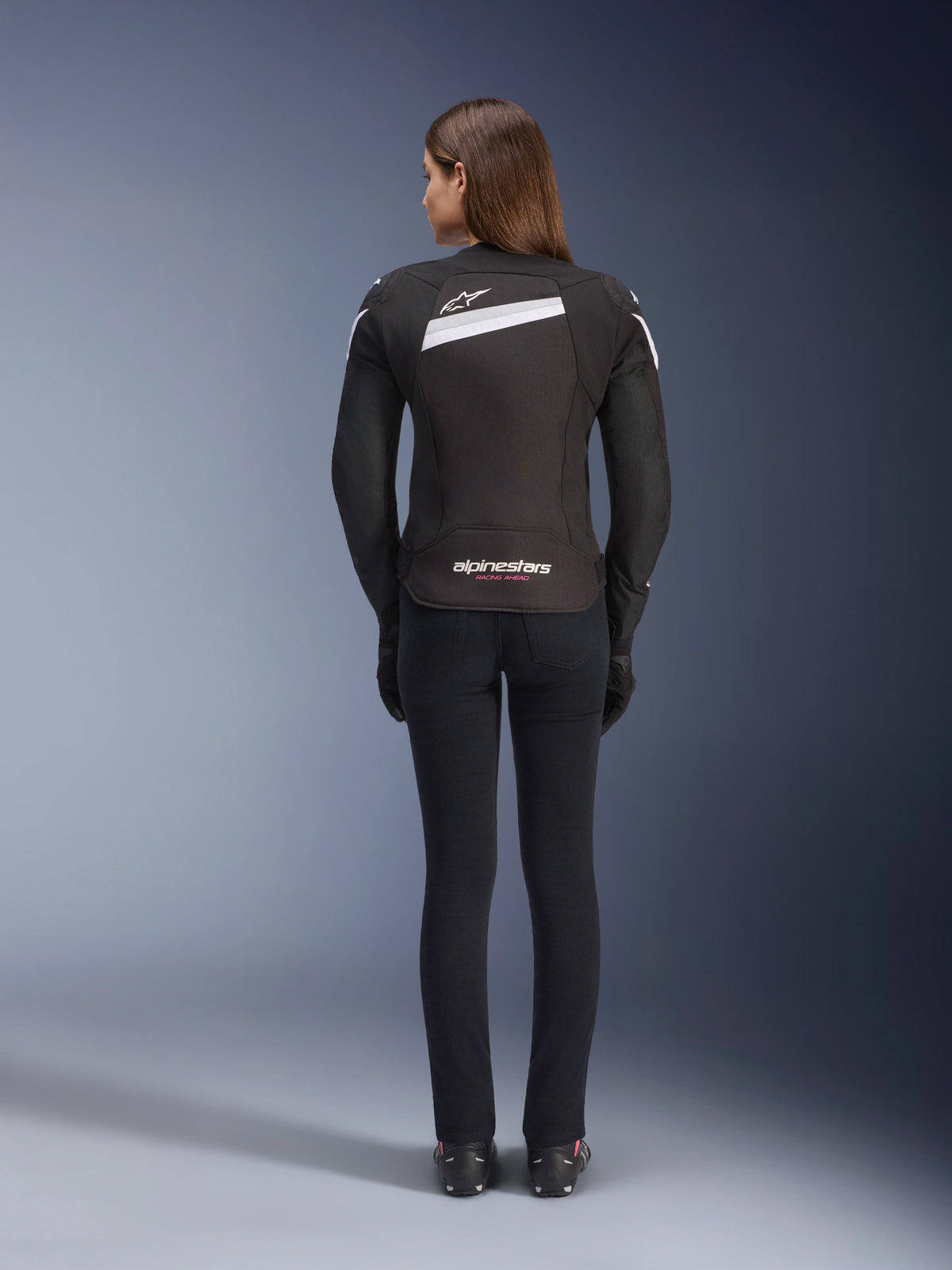 Veste Alpinestars Stella T-GP Plus R V4, veste de moto textile pour femme, noir et blanc, vue arrière montrant les accents blancs et le logo au dos, portée avec un pantalon noir, coupe sport profilée pour la performance sur route