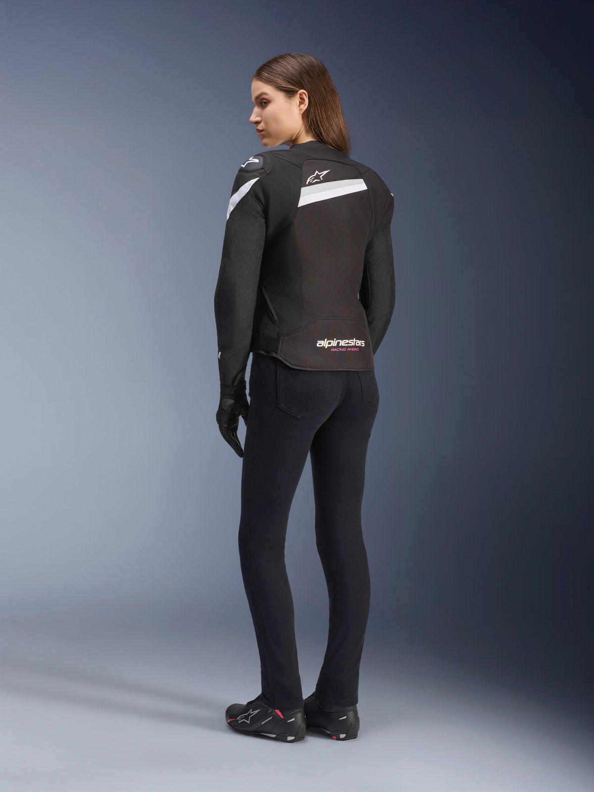 Veste Alpinestars Stella T-GP Plus R V4, veste de moto en textile pour femme, noir et blanc, vue de dos sur mannequin, avec accents blancs sur les épaules et logo Alpinestars, portée avec un pantalon noir et des gants de moto, coupe sport pour la conduite sur route