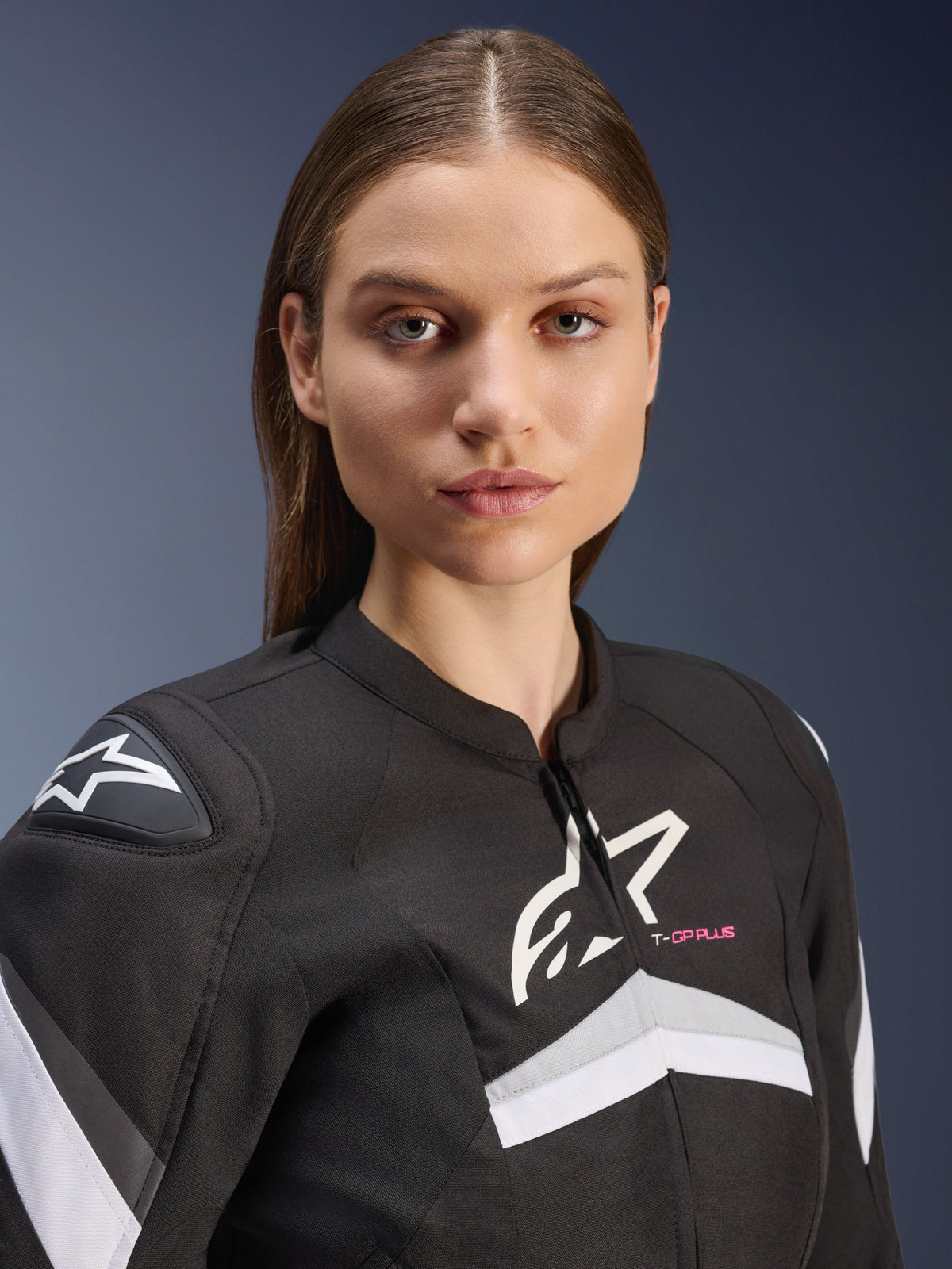 Veste Stella T-GP Plus R V4 Alpinestars, veste de moto en textile pour femme, noir et blanc avec accents gris et texte 'T-GP PLUS' rose, vue rapprochée d'un mannequin femme portant la veste à coupe sport avec logos Astars blancs