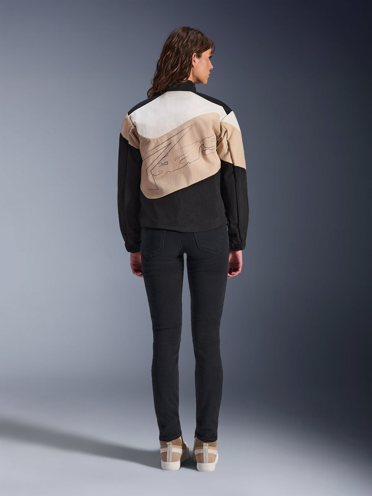 Veste Alpinestars Stella Moshell, veste de moto urbaine pour femme, vue de dos, design asymétrique noir, sable et blanc cassé avec un grand contour graphique, portée avec un pantalon slim noir et des baskets beiges, équipement de conduite urbaine