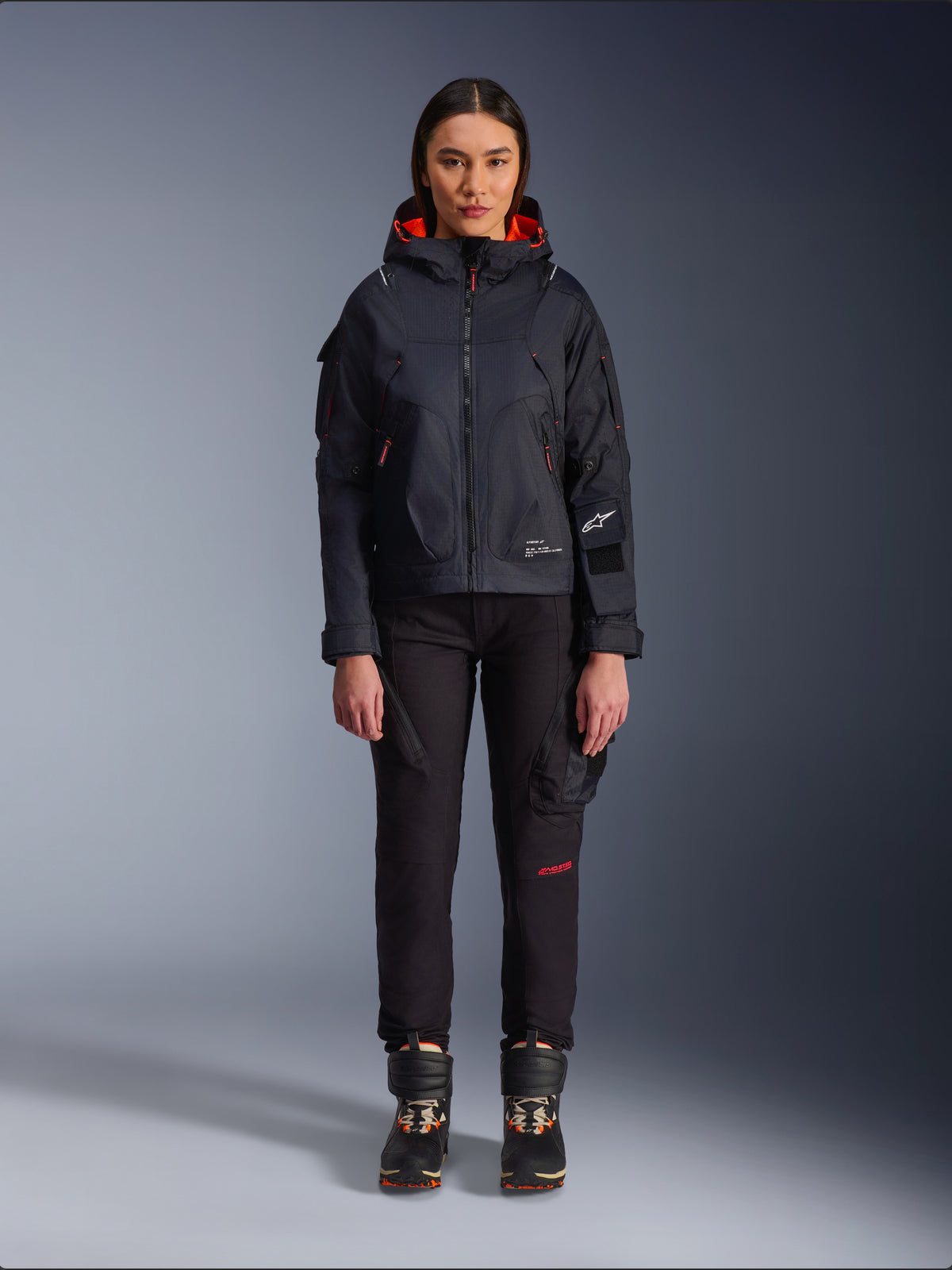 Alpinestars Actea Women's Pants, black motorcycle pants with red text detail and cargo pockets, worn by a female rider with a black hooded jacket, modern urban motorcycle apparel