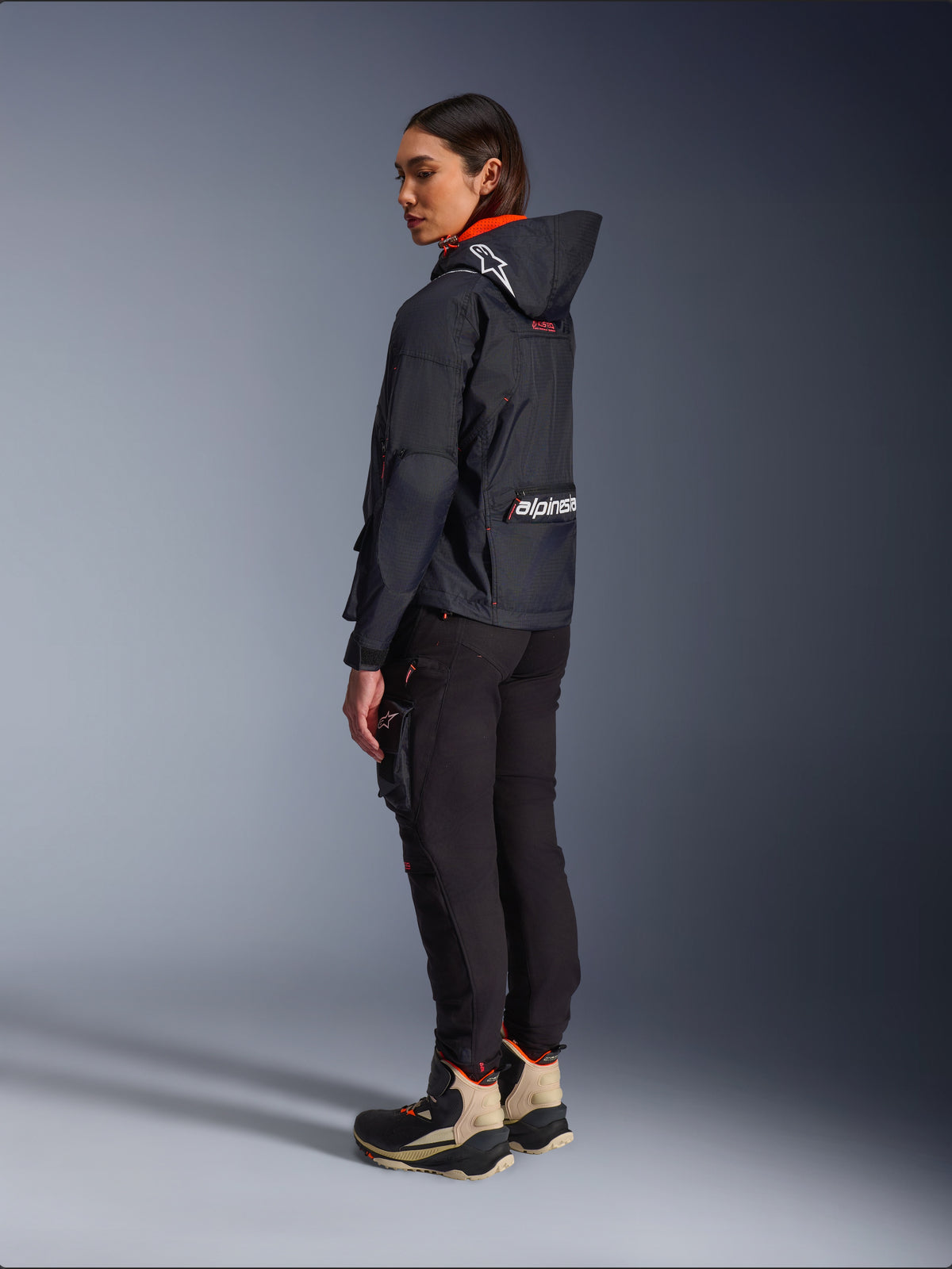 Motarde portant la veste Alpinestars Molly Air en Noir Corail Fluo, vue latérale arrière montrant la capuche avec logo Astars et grand marquage Alpinestars au dos, associée à un pantalon cargo de moto noir et des bottes d'aventure beiges et noires, équipement urbain contemporain pour femmes