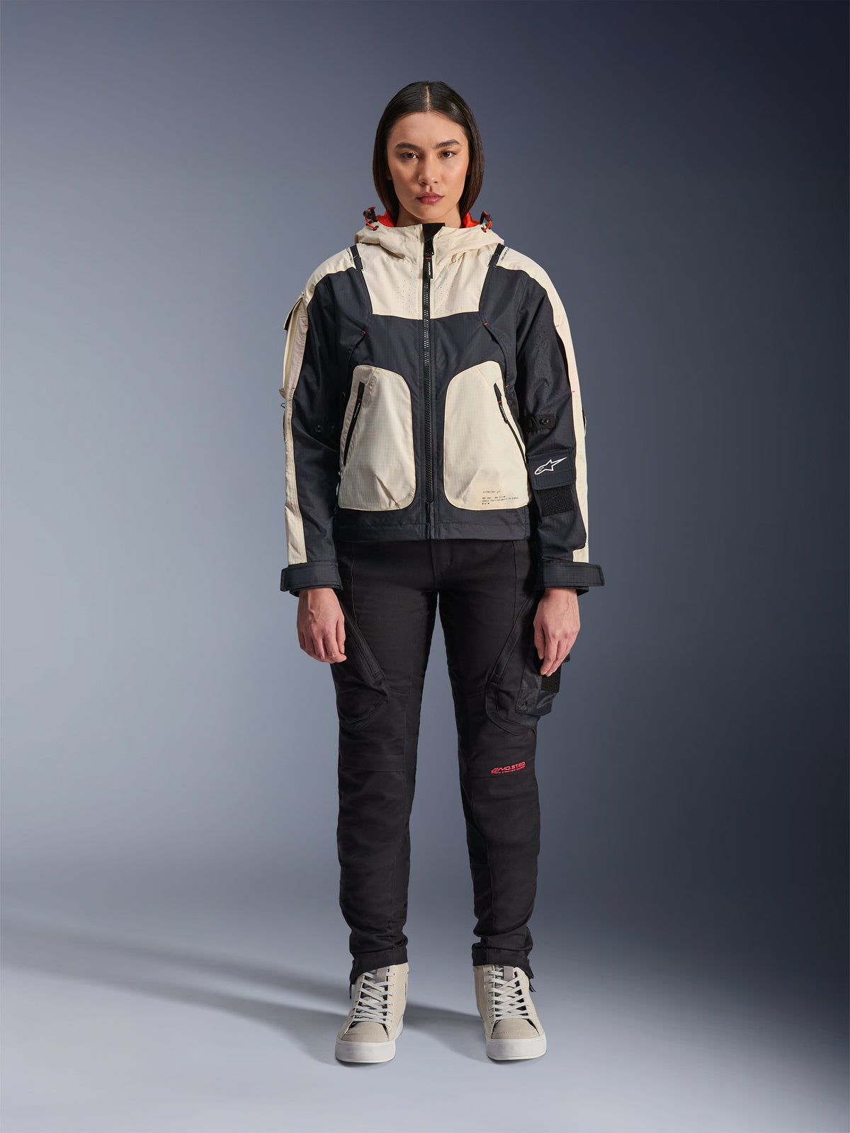 Mannequin femme portant la veste Alpinestars Molly Air en Black Almond, design color-block avec empiècements crème et noirs et capuche doublée de rouge, pantalon de moto noir et baskets crème, équipement de moto urbain contemporain, photo de studio