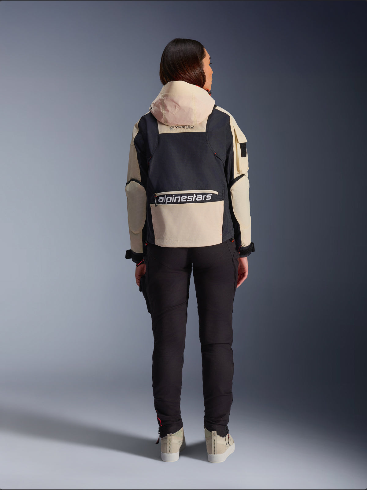 Veste Molly Air Alpinestars, veste de moto pour femme, Black Almond, vue arrière montrant la capuche et le grand logo au dos, design ripstop d'inspiration utilitaire, portée avec un pantalon de moto noir.
