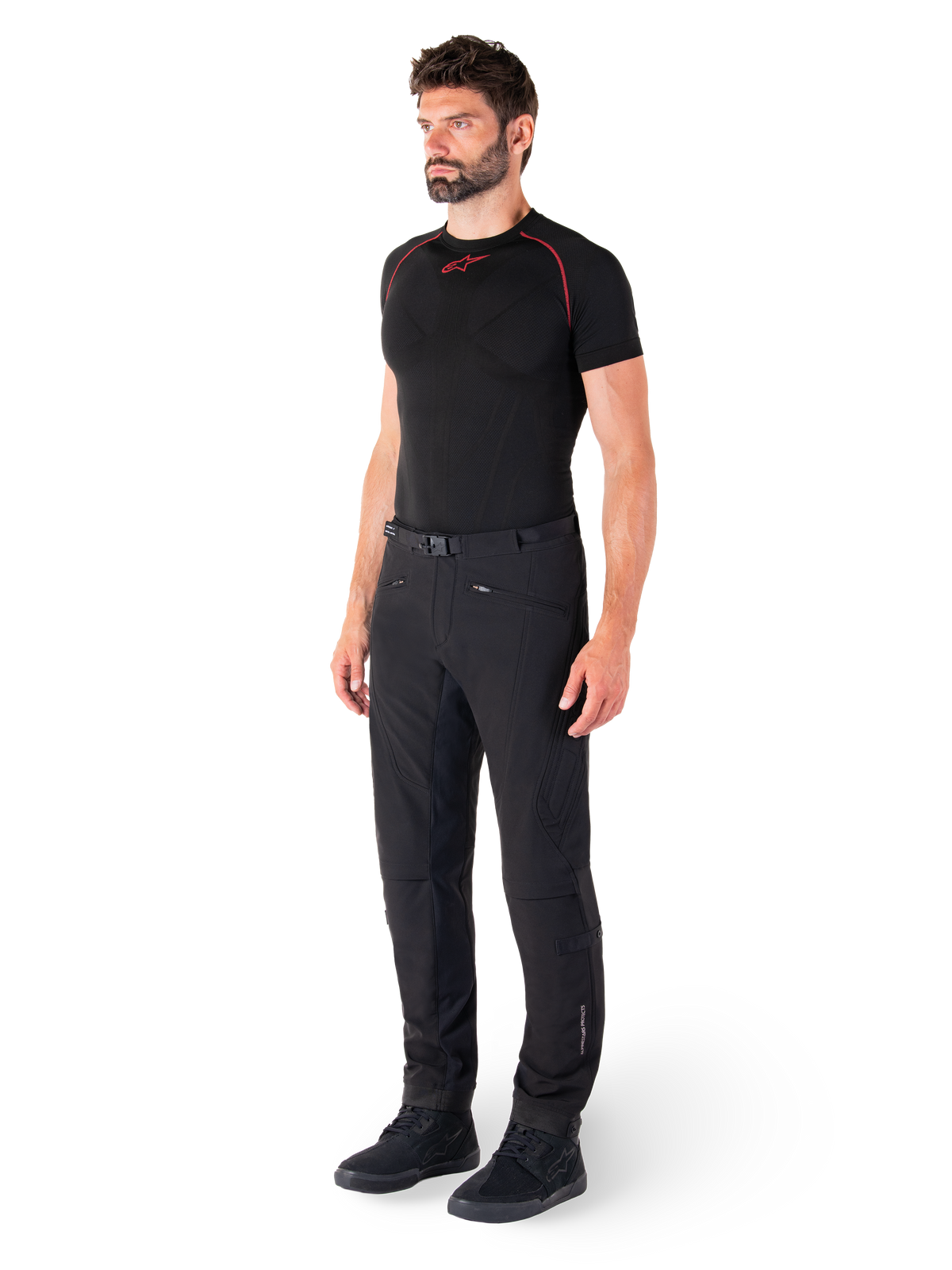 Pantalon Alpinestars Alden, pantalon de moto urbain, noir, textile softshell à coupe régulière, porté par un mannequin avec une couche de base noire et des chaussures de moto, doté d'une ceinture à fermeture magnétique et de poches zippées.