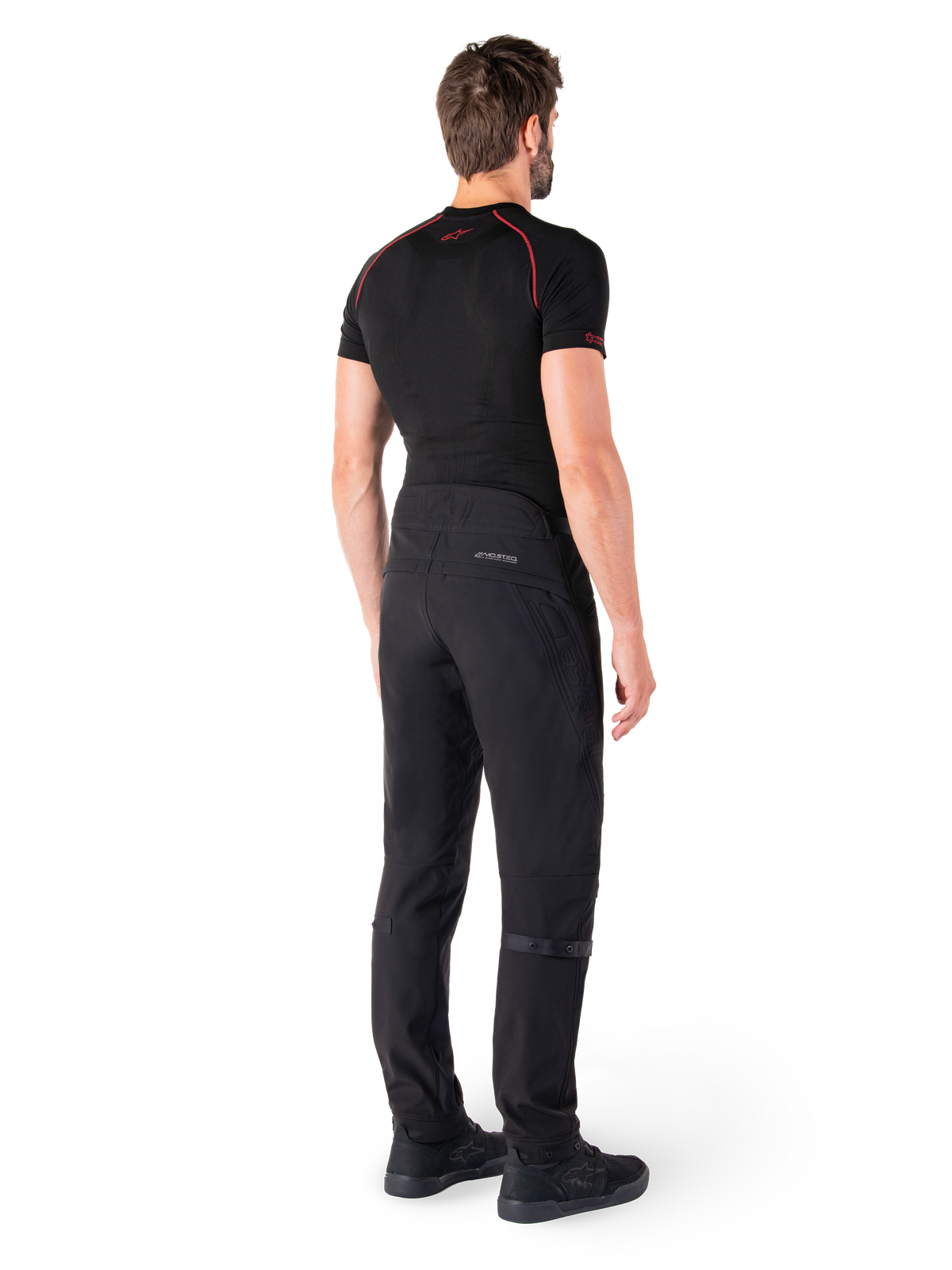Pantalon Alpinestars Alden, noir, pantalon de moto urbain présenté de dos sur un mannequin, porté avec une couche de base noire à manches courtes et des chaussures de moto noires, avec des détails matelassés et le marquage réfléchissant MO.ST.EQ. sur l'empiècement arrière.