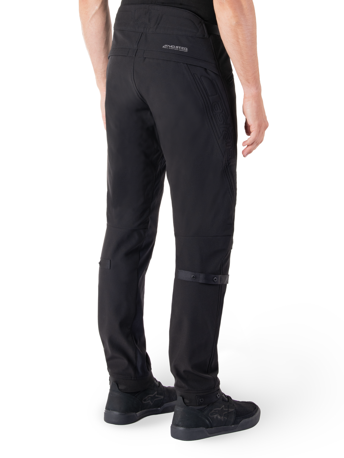 Pantalon Alpinestars Alden, pantalon de moto urbain, noir, vue arrière montrant le tissu softshell technique avec des détails matelassés, marquage réfléchissant MO.ST.EQ. sur l'empiècement arrière et sangles de mollet réglables, associé à des chaussures de moto noires