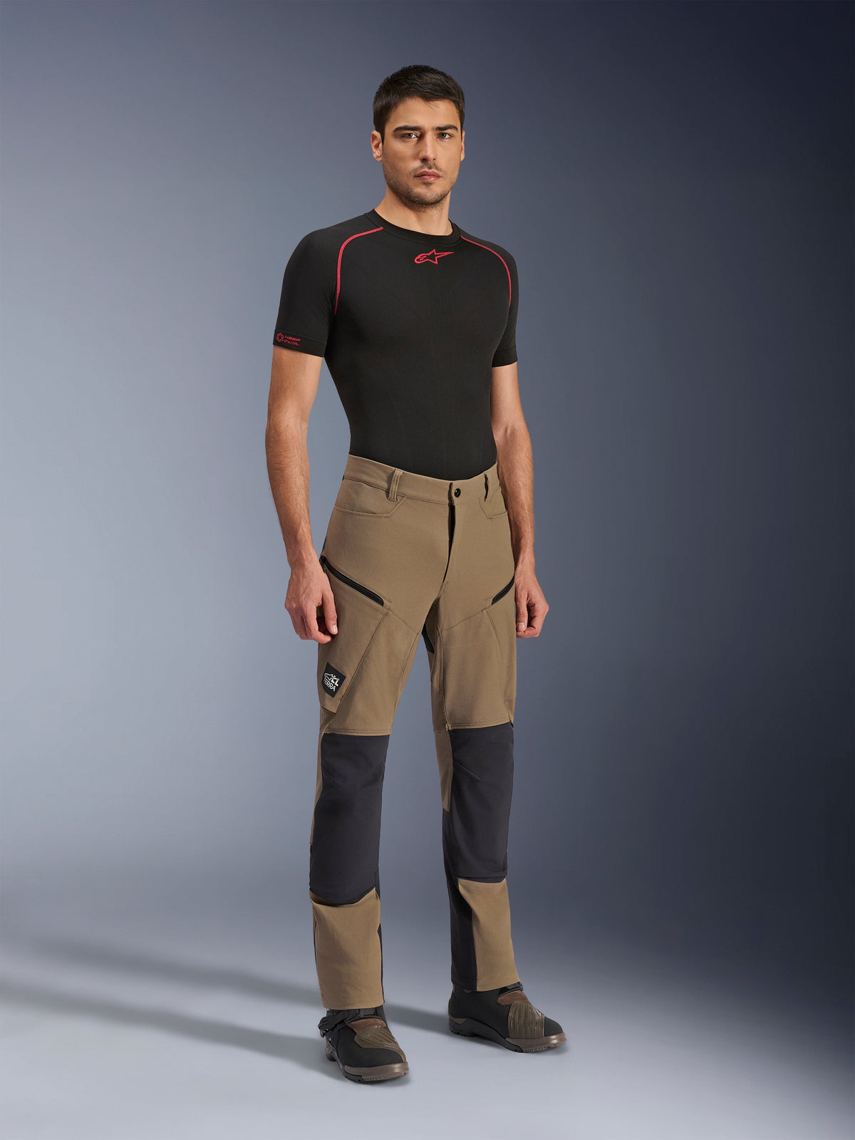 Alpinestars Flex-AST Explorer Pants, Walnut Black adventure motorcycle pants, worn by a male model with a black short-sleeve base layer and adventure boots, featuring zippered cargo pockets and reinforced knee panels, technical gear for dual-sport and touring riders.