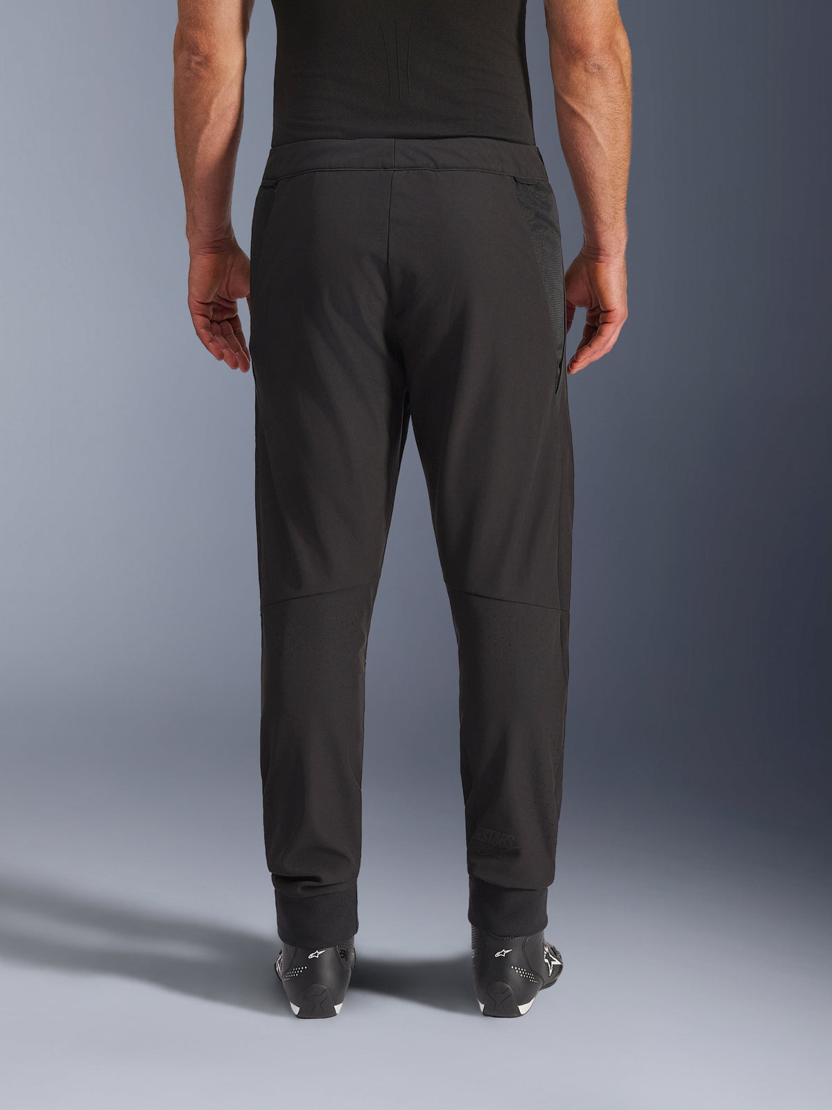 Pantalon Alpinestars Aeroshell Airflow, noir, vue arrière, doté de zones perforées au laser sur les mollets et de panneaux en maille, porté avec des bottes Alpinestars noires, conception softshell légère pour le sport et le touring