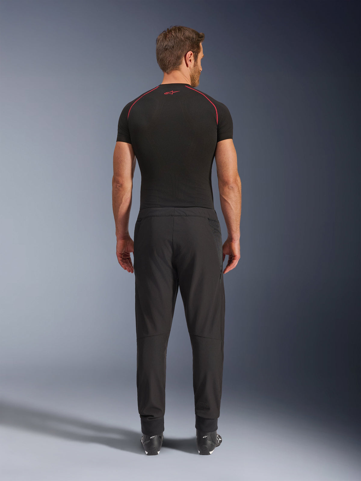 Pantalon Alpinestars Aeroshell Airflow, pantalon de moto noir, vue de dos sur un mannequin avec une couche de base noire et des bottes, doté d'un châssis en softshell léger avec des panneaux en maille intégrés pour la ventilation.