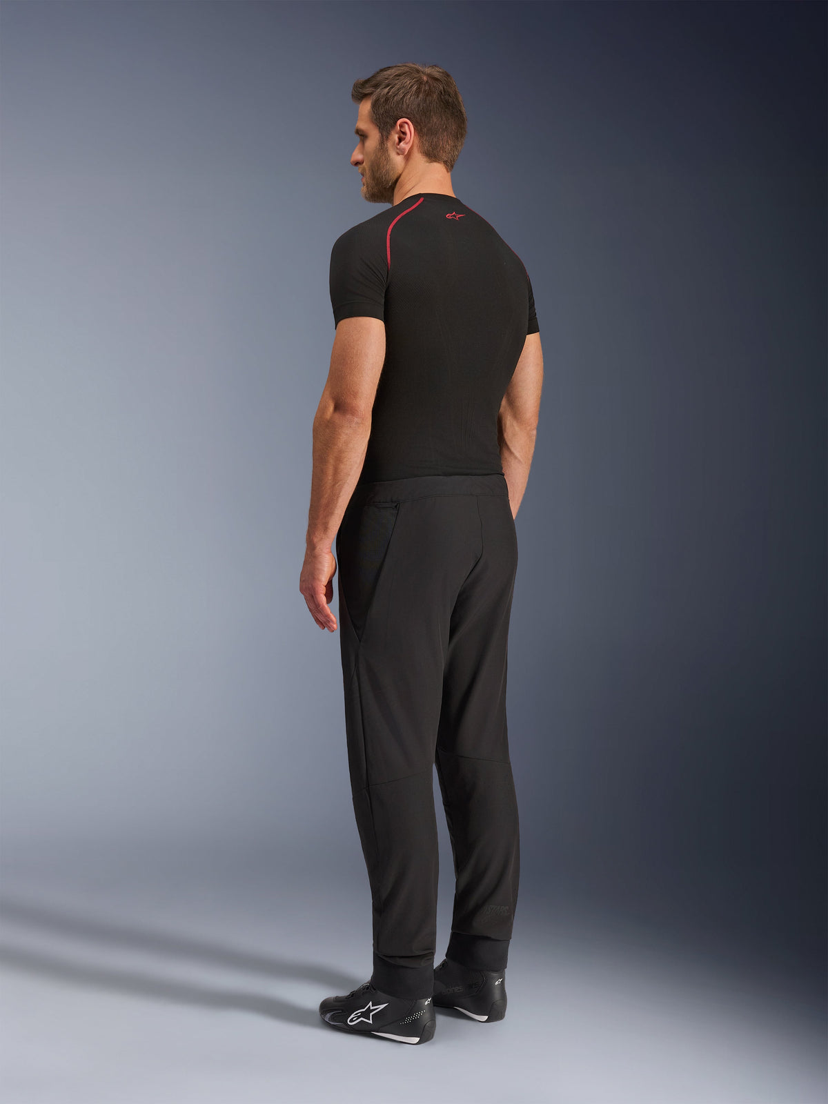 Pantalon Alpinestars Aeroshell Airflow, pantalon de moto noir, textile softshell léger, porté avec un t-shirt technique noir à manches courtes et des bottes de moto noires, vue de profil arrière montrant la coupe ajustée et la construction ergonomique.