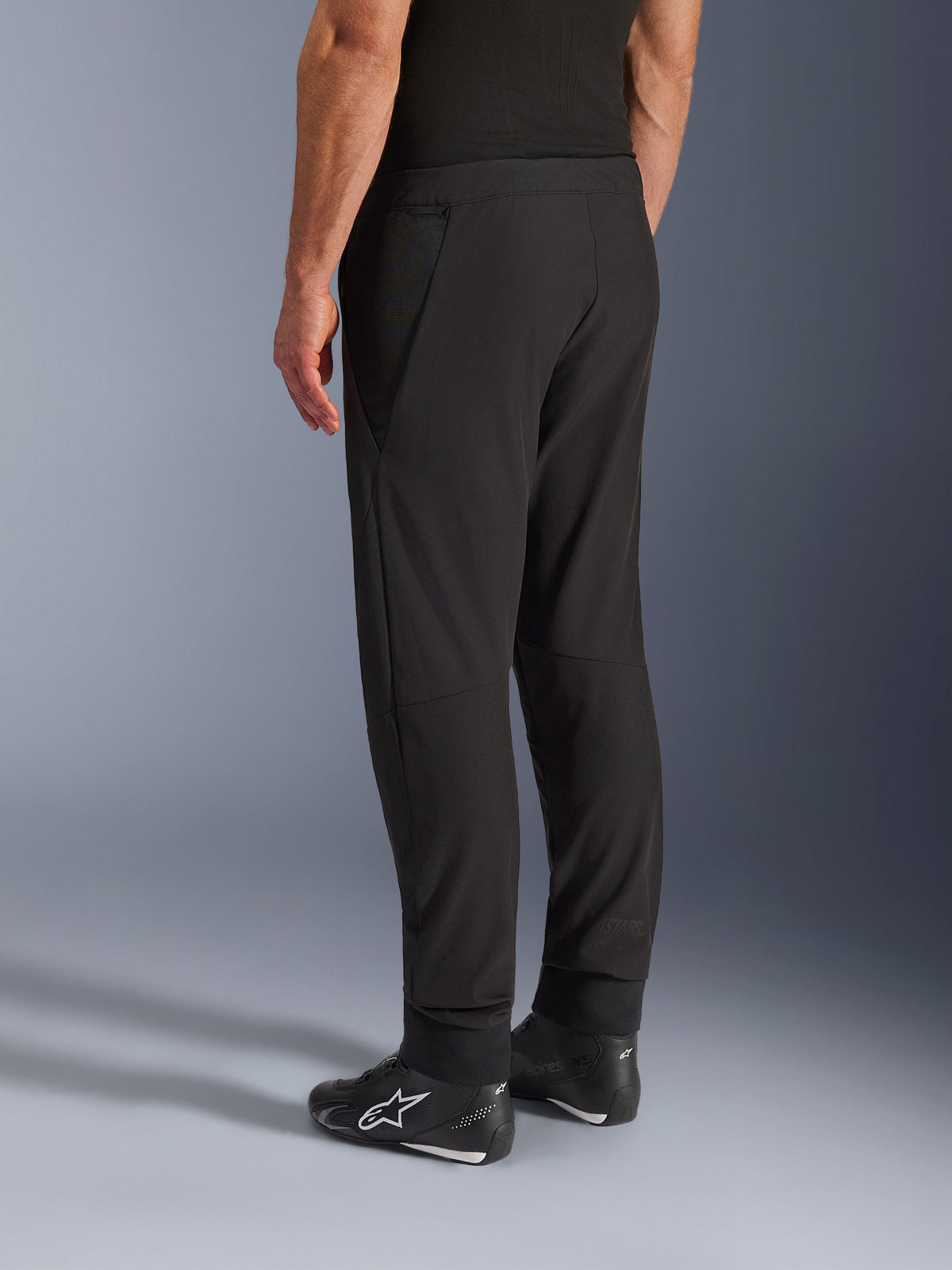 Pantalon Alpinestars Aeroshell Airflow, pantalon de moto noir, vue arrière de trois quarts, construction en softshell extensible légère avec panneaux en maille intégrés, poignets de cheville réglables, porté avec des chaussures de moto Alpinestars noires