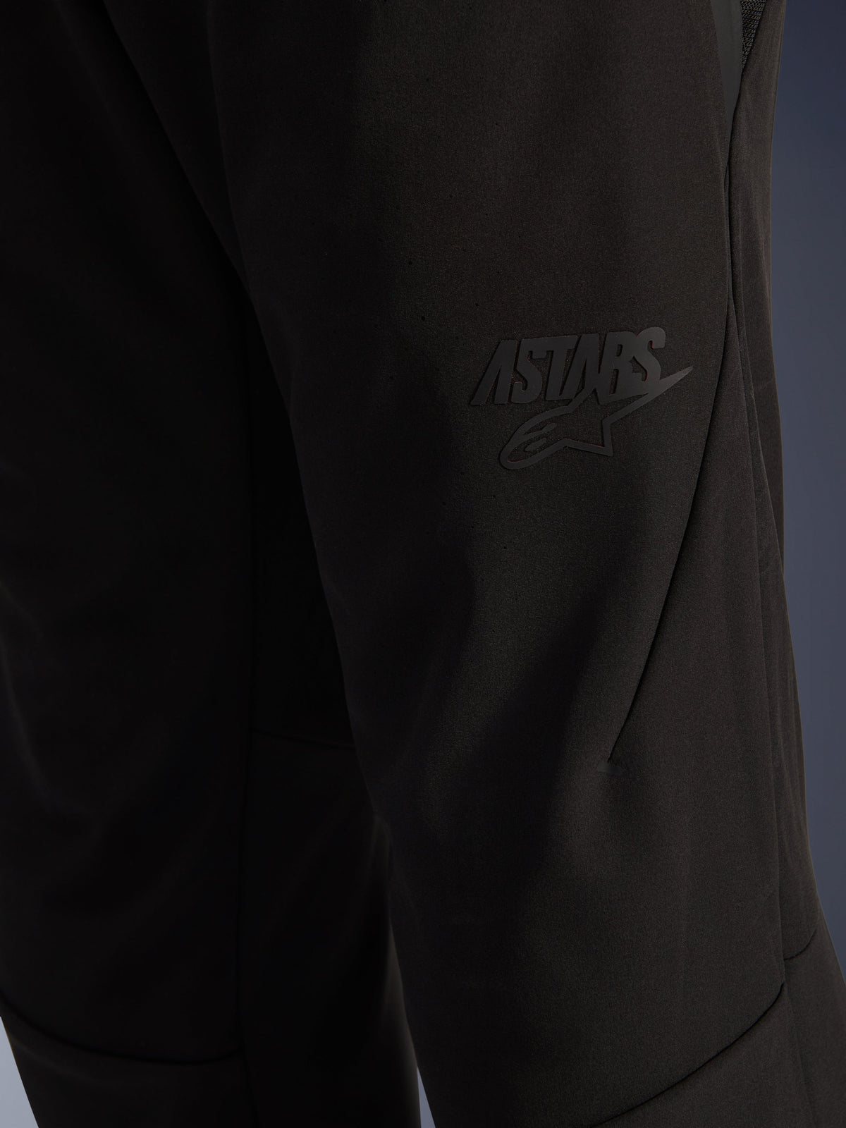 Pantalon Alpinestars Aeroshell Airflow, pantalon de moto noir, vue rapprochée de la cuisse avec logo ASTARS noir en relief, matériau softshell extensible léger et coutures ergonomiques