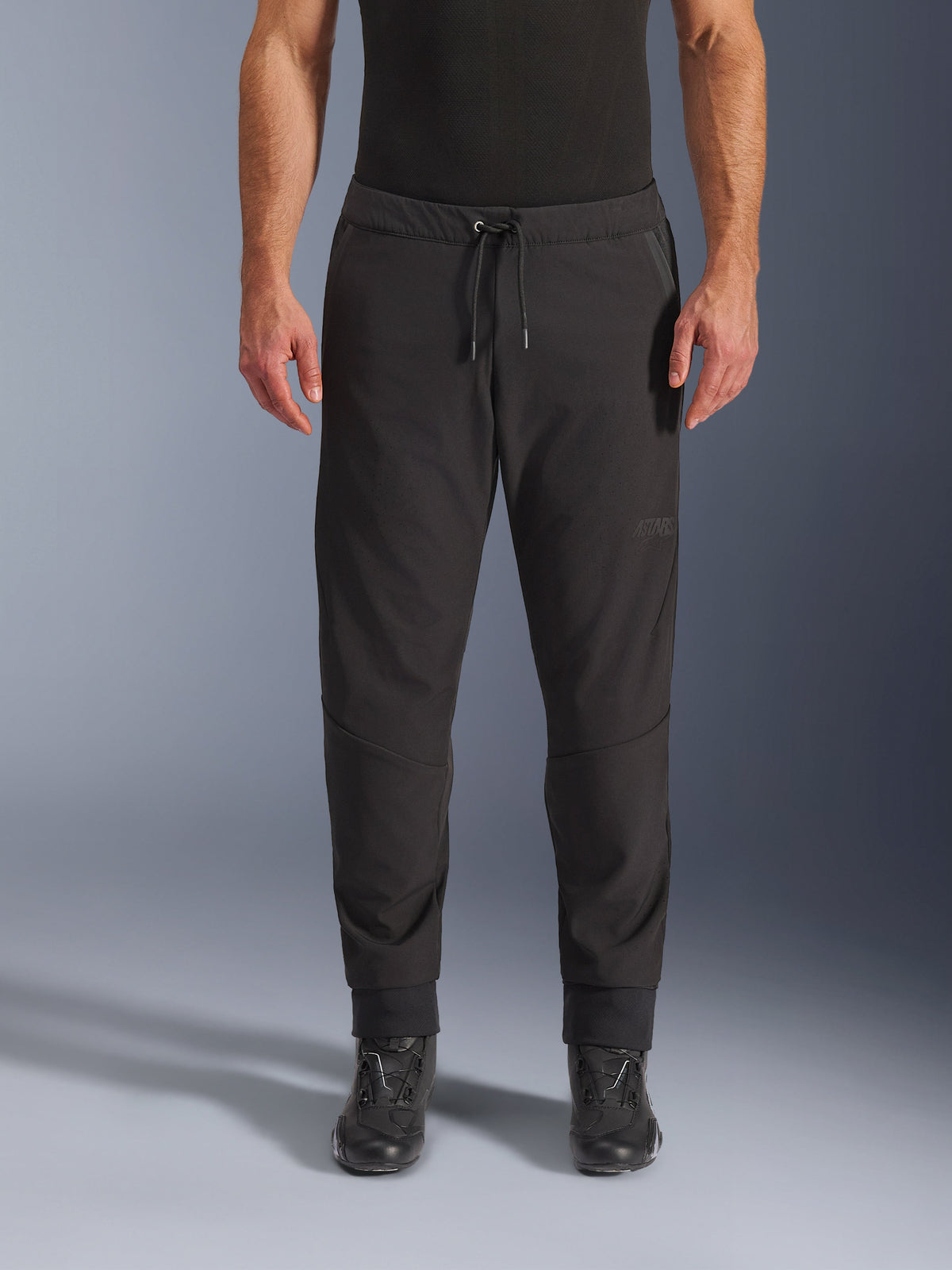 Alpinestars Aeroshell Airflow Pants, pantalon de moto noir, vue de face porté avec des bottes de moto noires, doté d'une taille à cordon de serrage et de poignets de cheville fuselés, design en softshell léger avec logo ton sur ton discret.