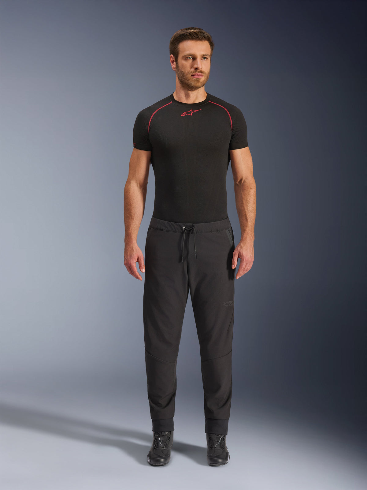 Modèle portant le pantalon Alpinestars Aeroshell Airflow en noir, associé à une couche de base noire à manches courtes et des bottes de moto noires. Ce pantalon de moto en softshell léger présente une taille à cordon de serrage et des poignets élastiques, conçu pour les motards de sport et de tourisme par temps chaud.